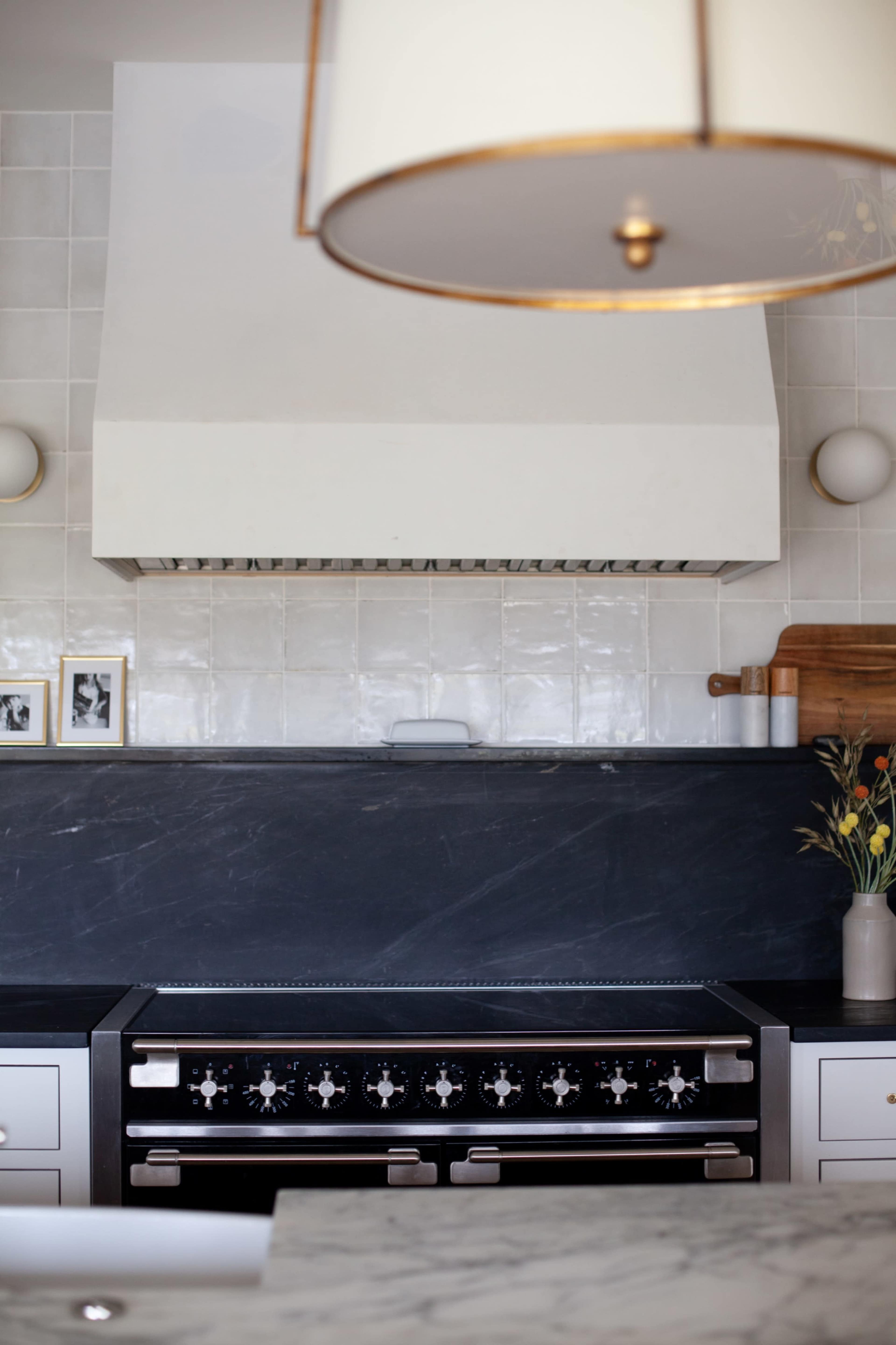 The image shows a modern kitchen with a stainless steel range, a hood ventilator, and a dark stone backsplash, complemented by a simple light fixture overhead.