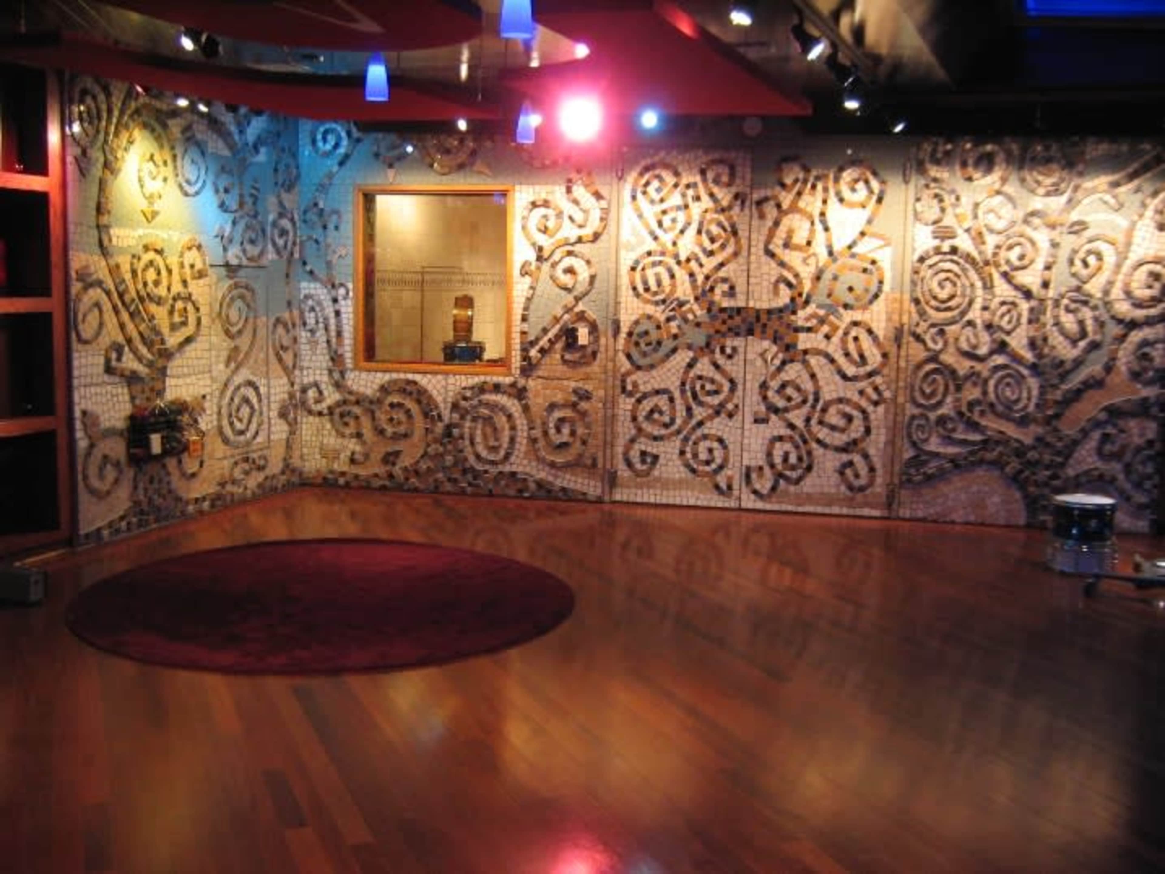 The image shows a gallery space with a circular red rug on a wooden floor, featuring intricate mosaic designs on the walls and blue lights hanging from the ceiling.