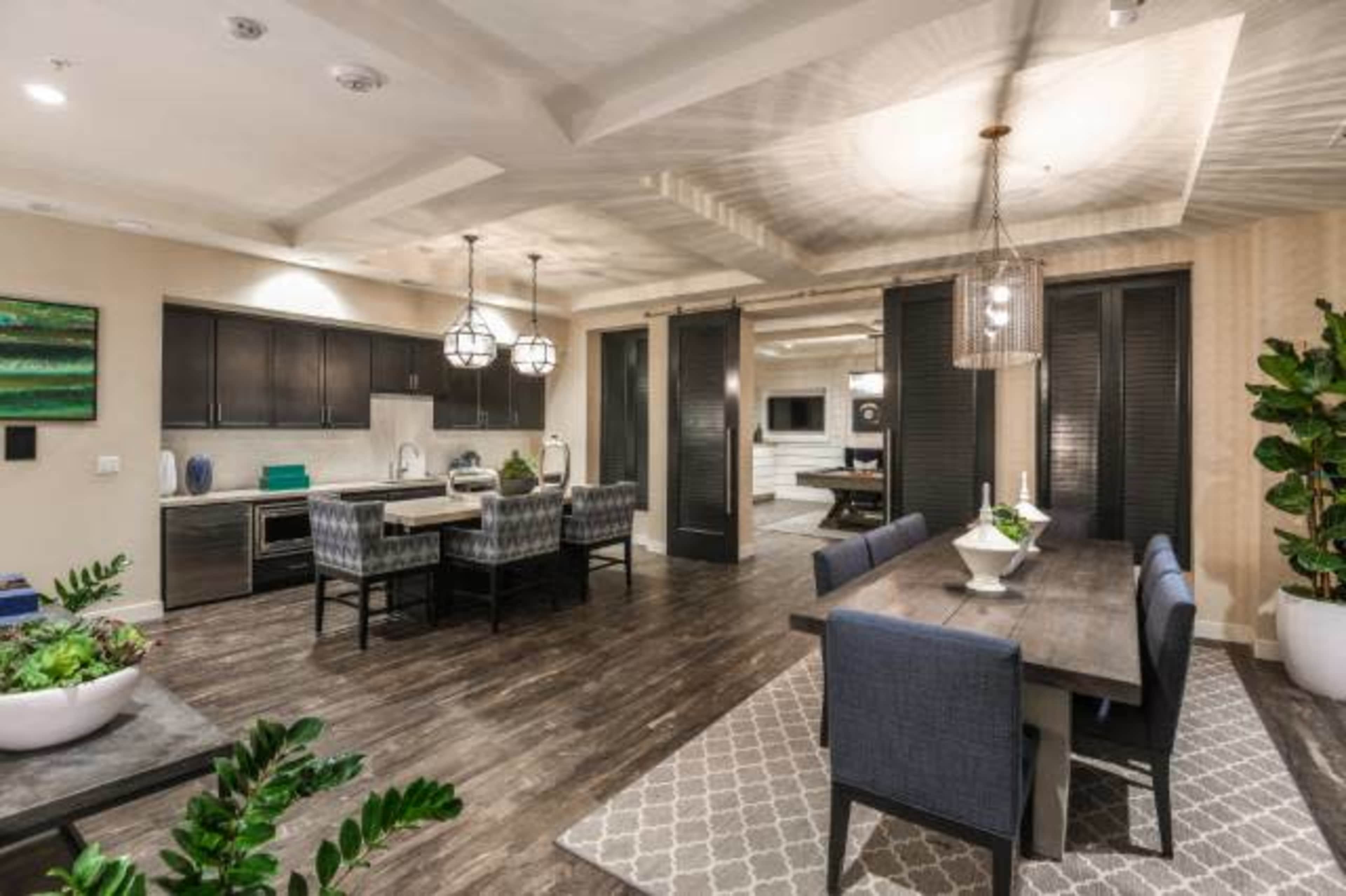 An open-concept dining and kitchen area featuring a wooden dining table, upholstered chairs, modern light fixtures, and plants.