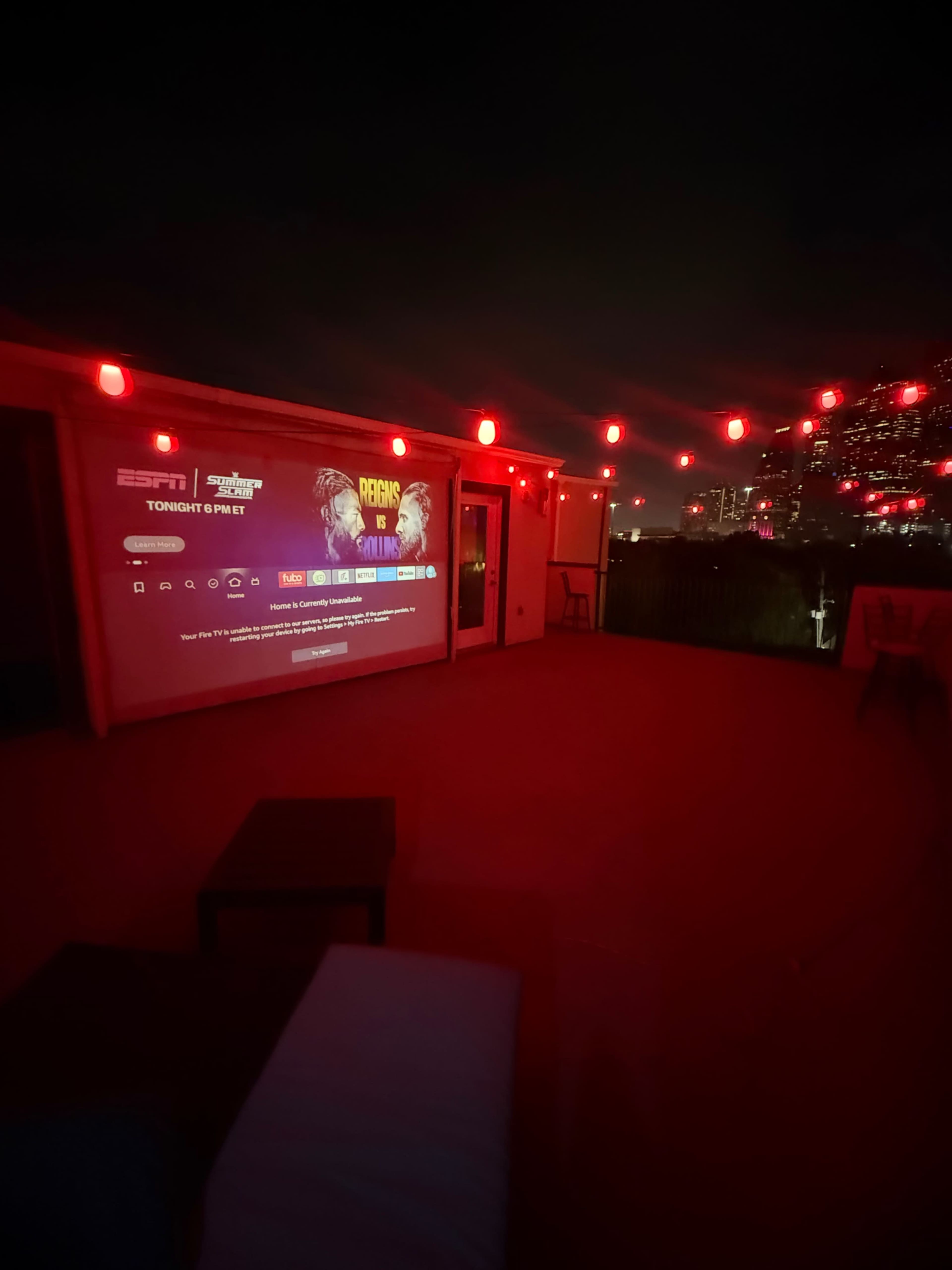 The image shows a rooftop setting illuminated by red lights, featuring a wall projection of a sports program alongside a city skyline at night.