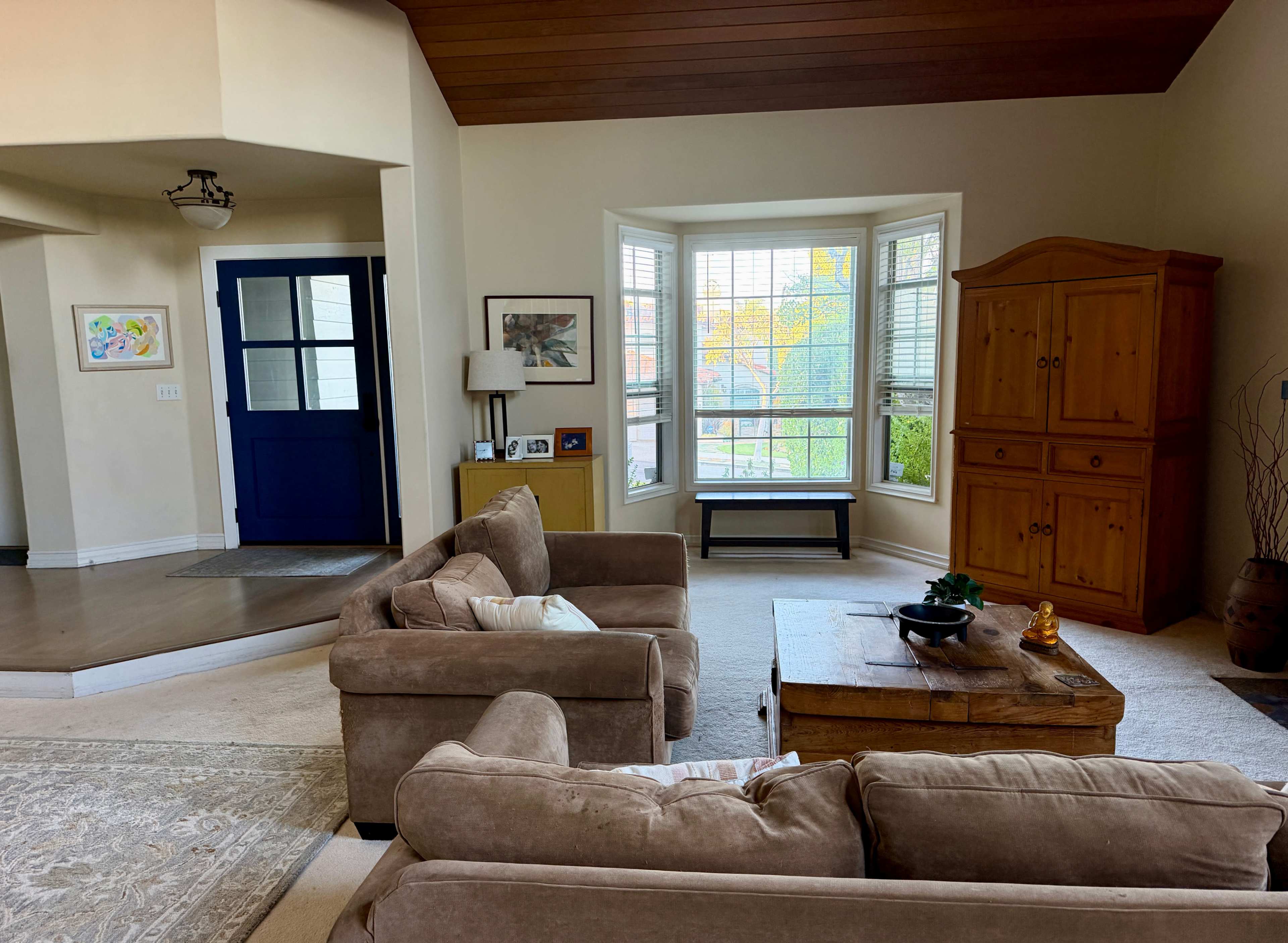 The image shows a living room with beige sofas, a wooden coffee table, and a bay window that lets in natural light, while a blue door leads to another area.