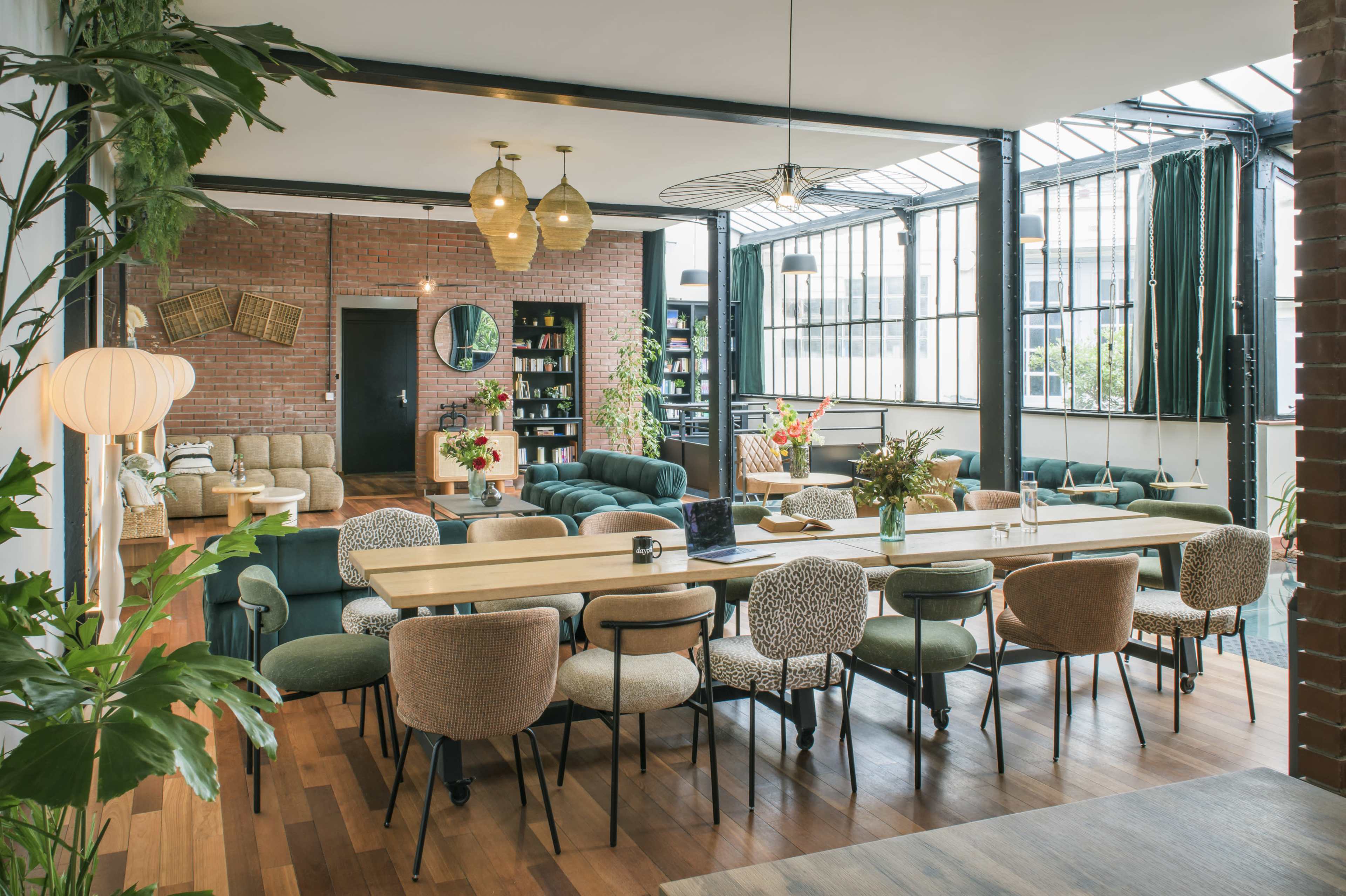 The image features a modern, open-concept interior of a lounge or dining area with a large wooden table surrounded by various chairs, lush greenery, and a mix of seating arrangements.