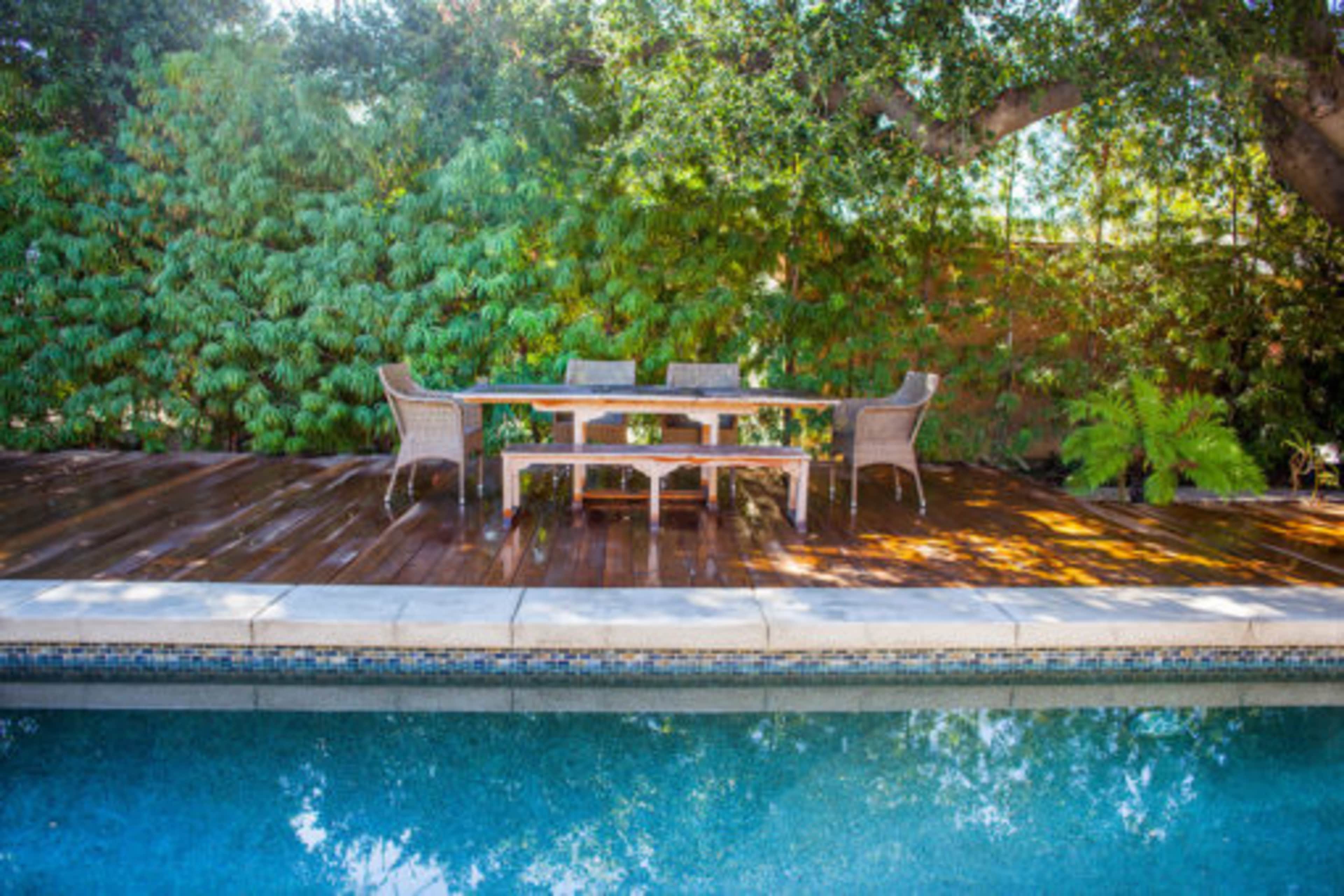 A wooden deck with a dining table and chairs is positioned next to a swimming pool, surrounded by lush greenery.