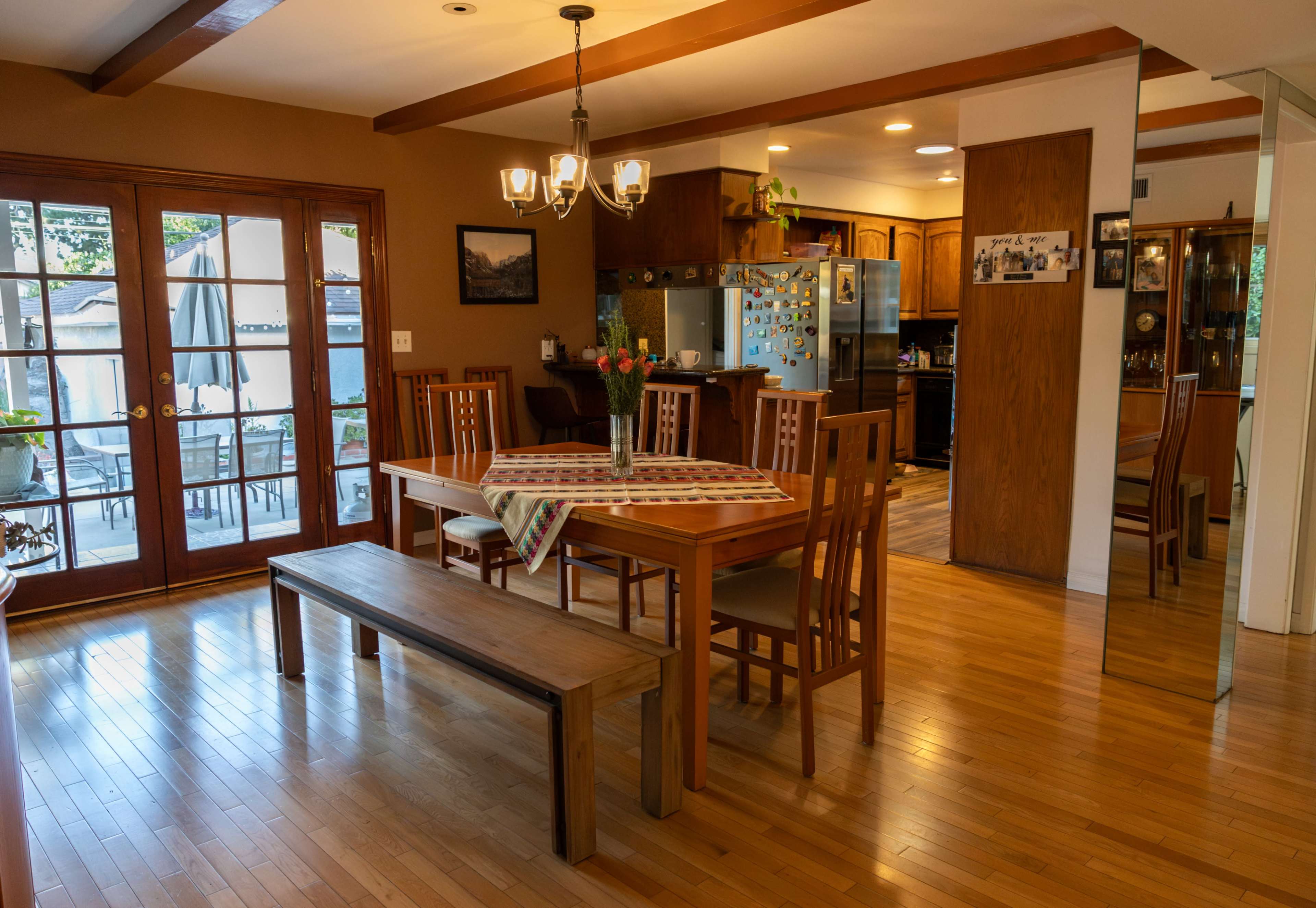 A wooden dining table with chairs in a bright room that opens to a patio with sliding glass doors, and a kitchen area in the background.