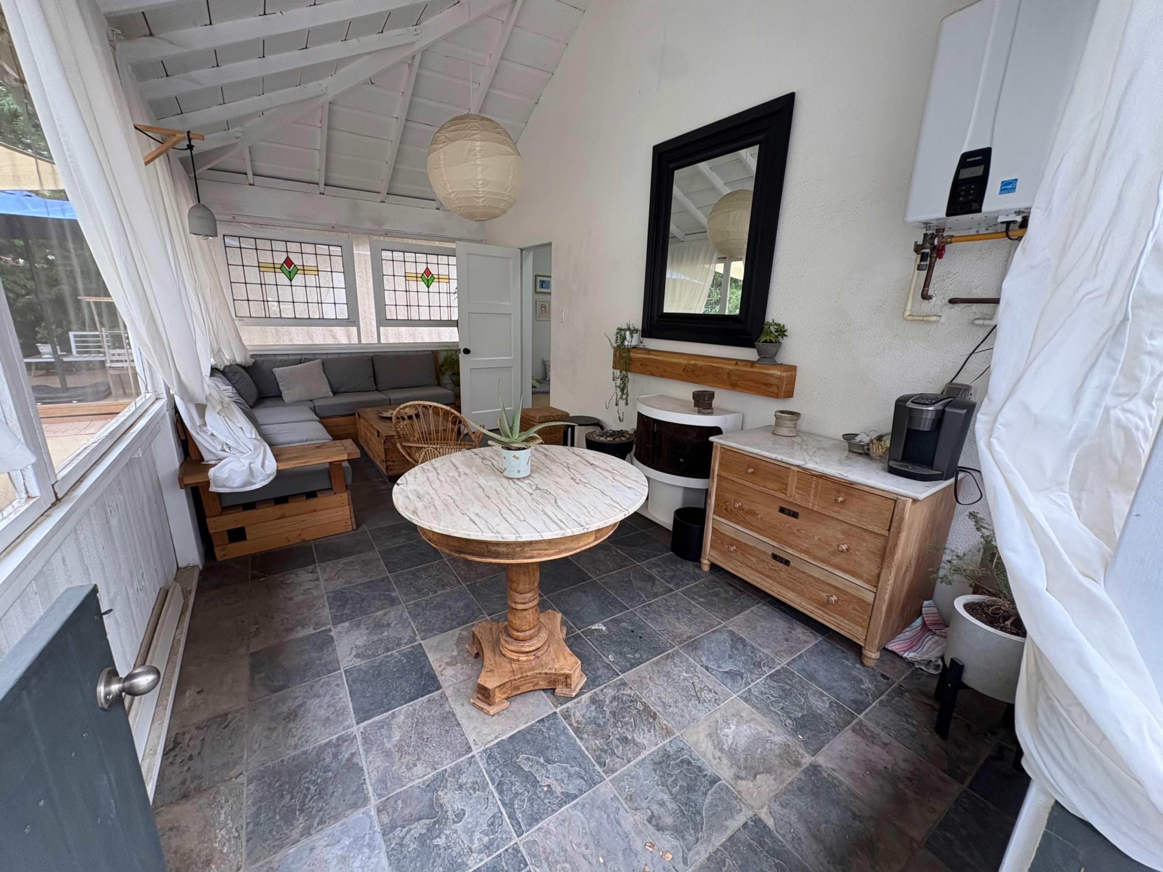 The image shows a cozy indoor space with a round table, wooden chairs, a sofa, and various decor items against a light-colored wall.