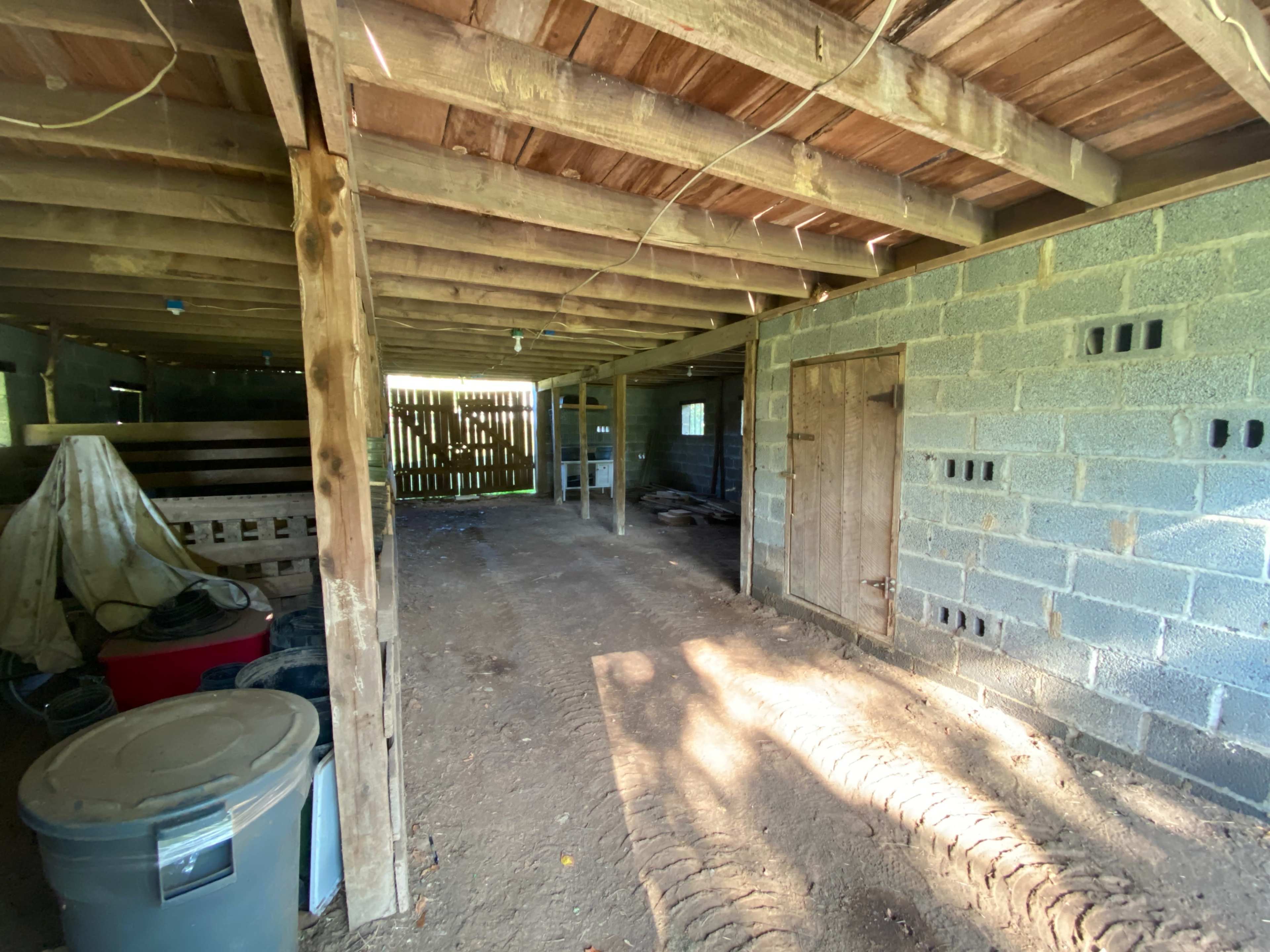 The interior of a rustic barn with exposed wooden beams, concrete walls, and dirt flooring, featuring an empty space on one side and a wooden door on the other.