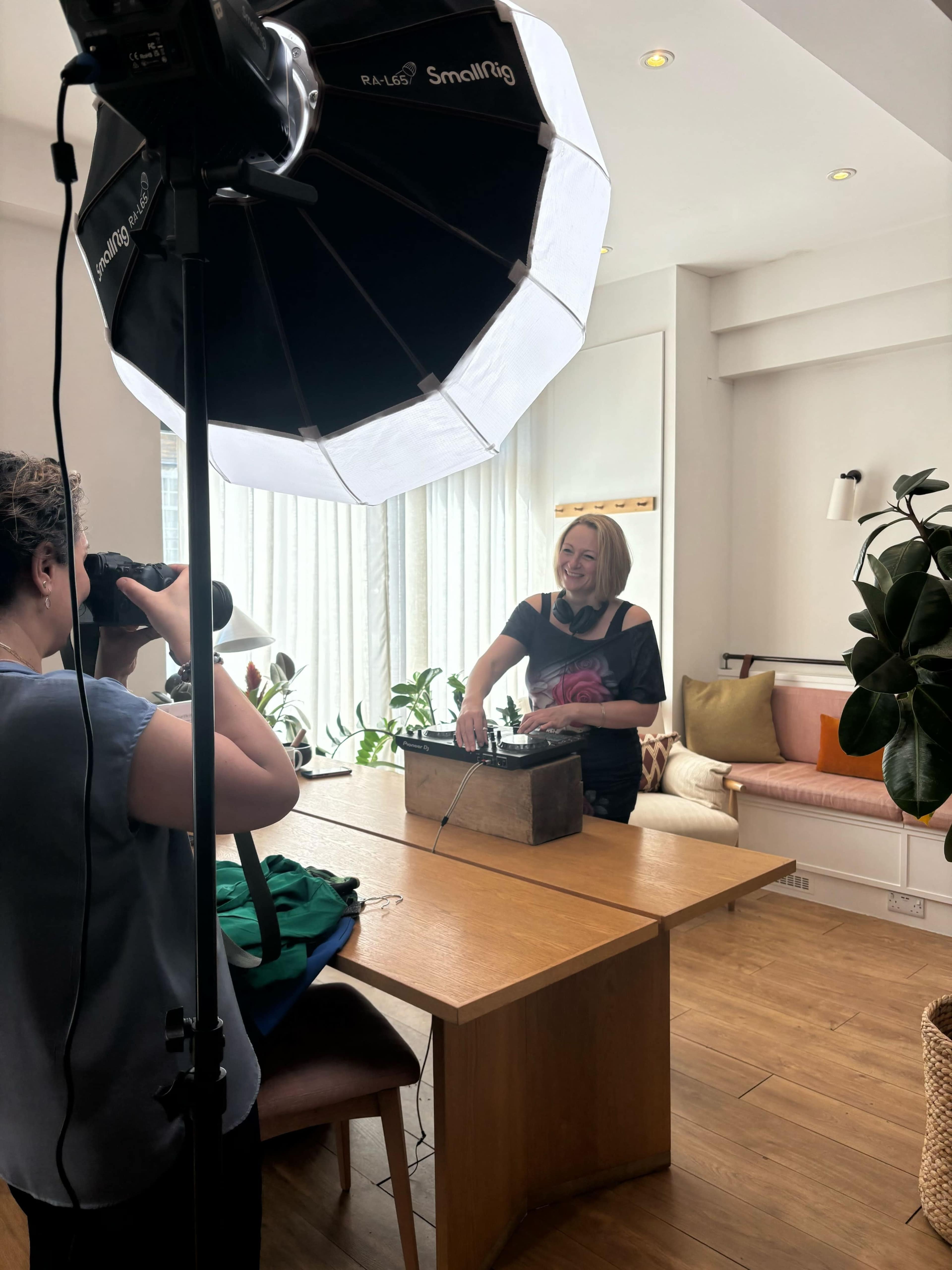 A woman stands behind a wooden table with a vintage item while another person takes her photo using a camera with a large softbox light.
