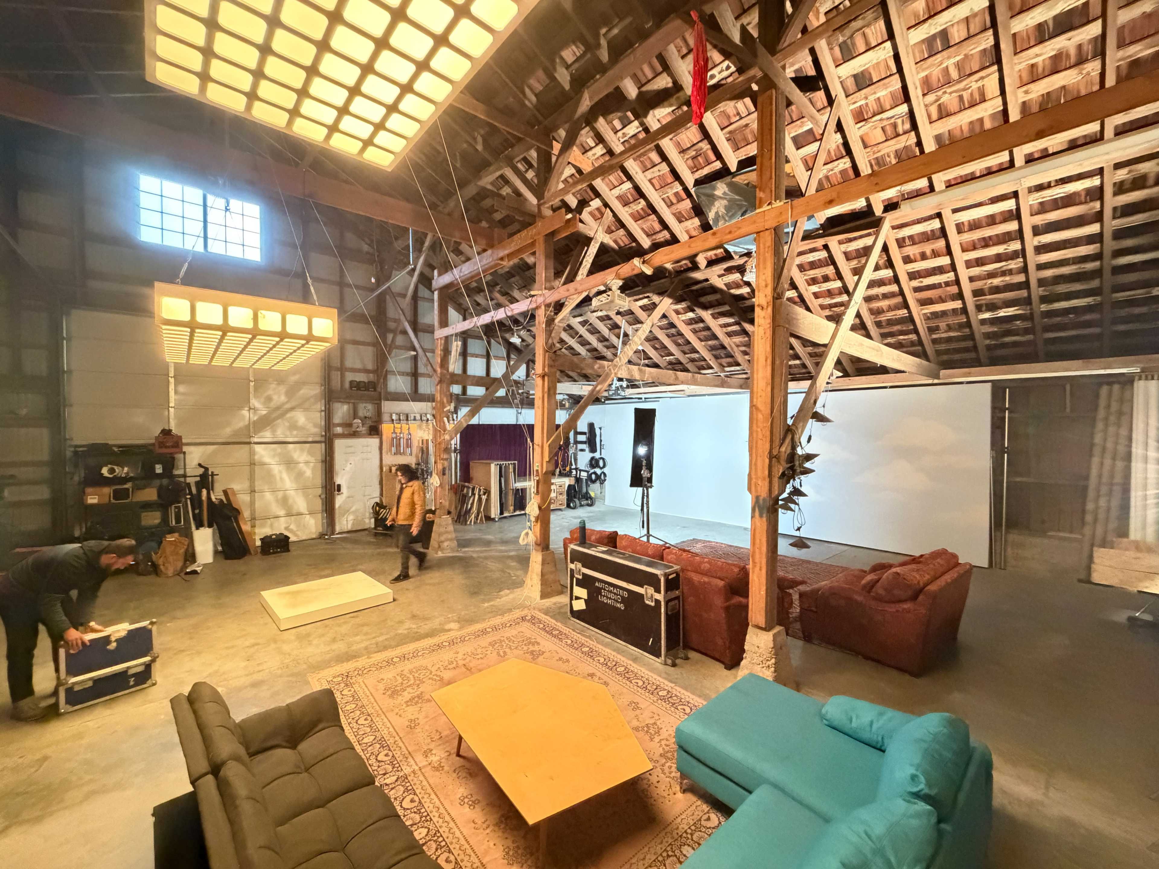 The image shows a large, open barn interior featuring a stage, various seating arrangements with sofas, and equipment for sound and lighting.