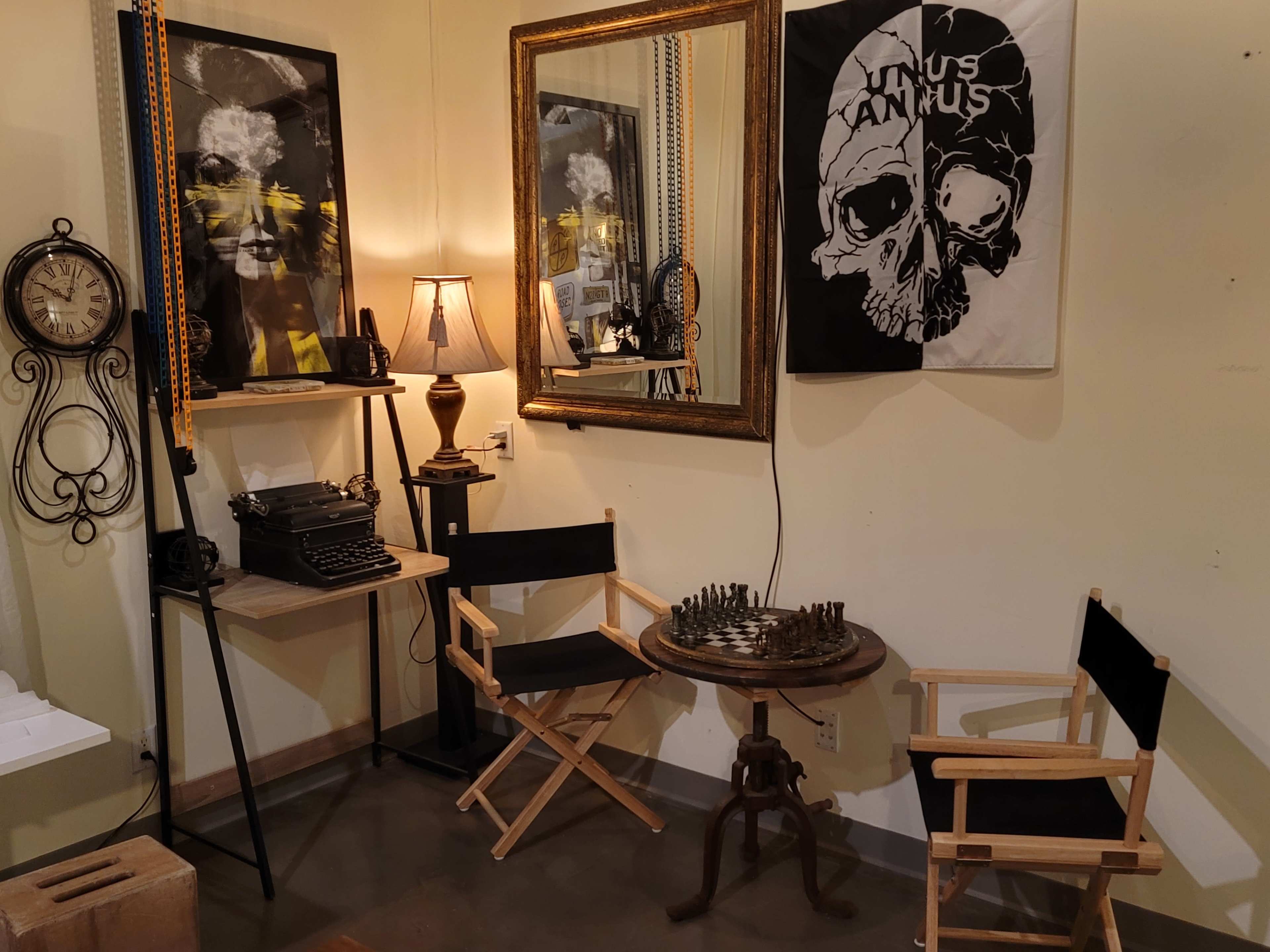 The image features a small room with a vintage typewriter, a chess set, two director's chairs, a lamp, a large mirror, and artwork on the wall, including a flag with a skull design.