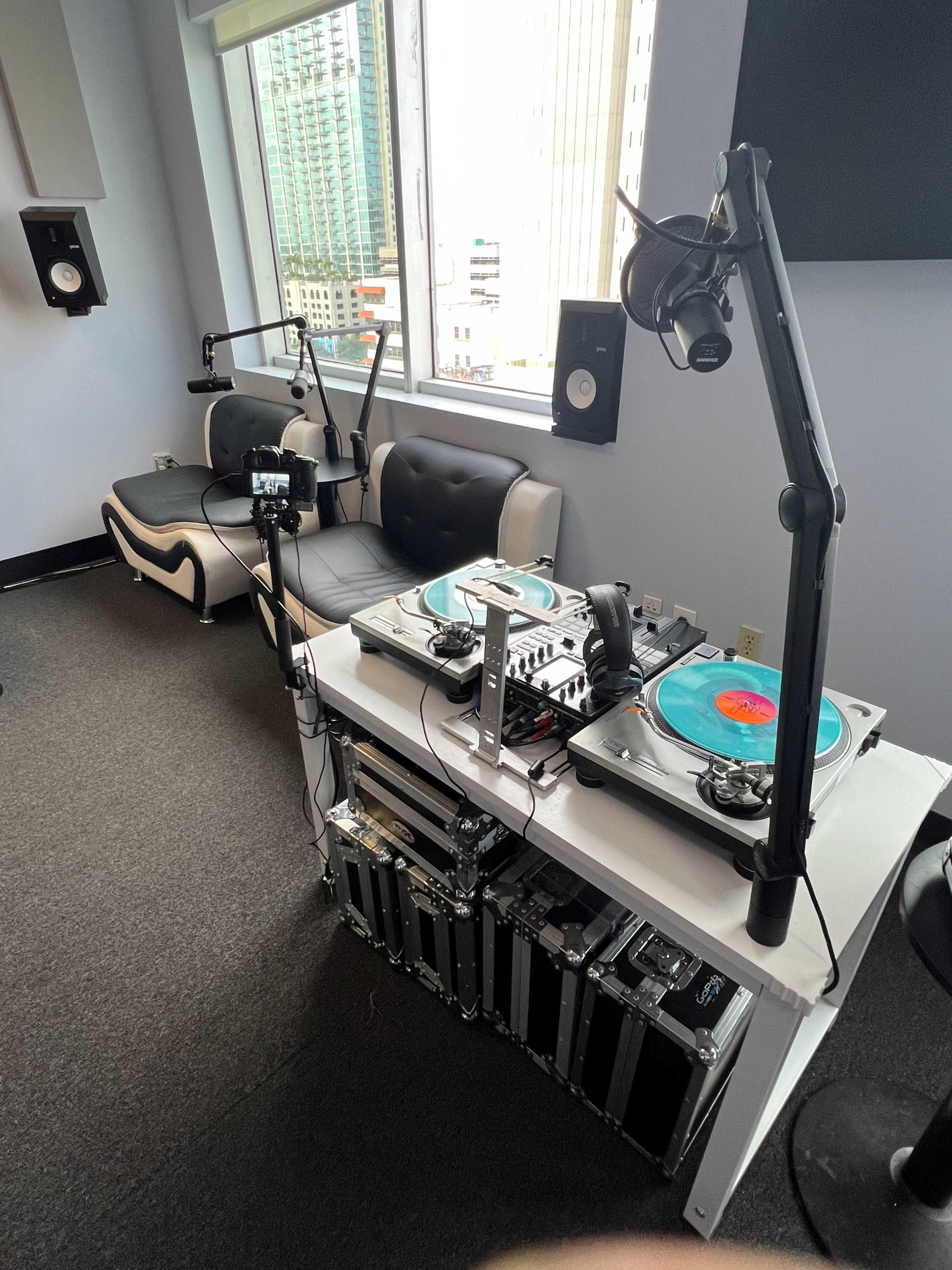 A DJ booth featuring turntables, audio equipment, and microphones is set up next to a seating area in a room with large windows.