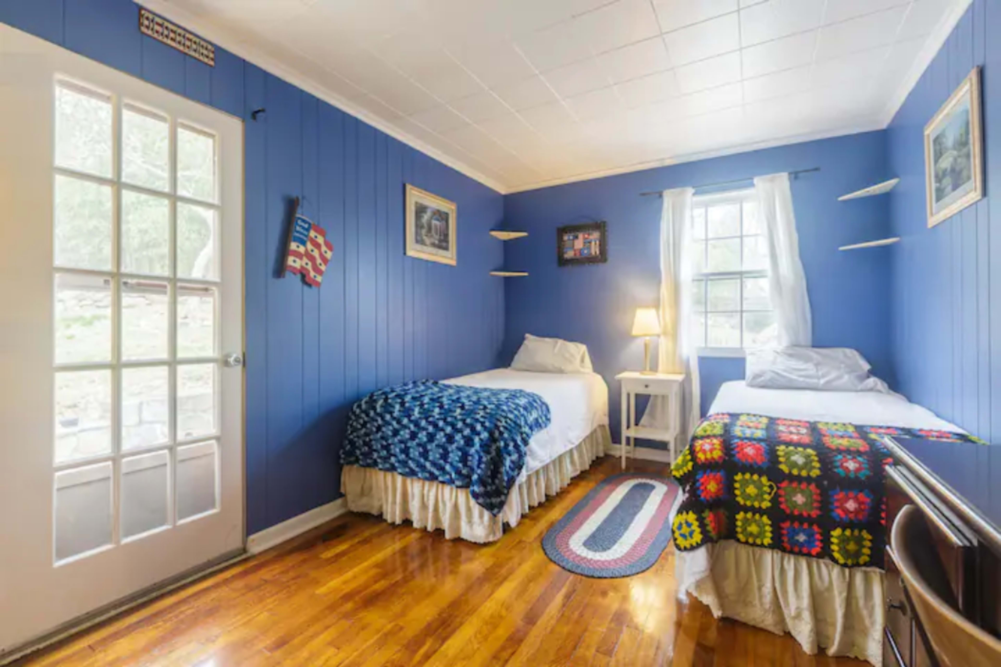 A blue-walled bedroom with two twin beds, a small nightstand with a lamp, and hardwood floors.