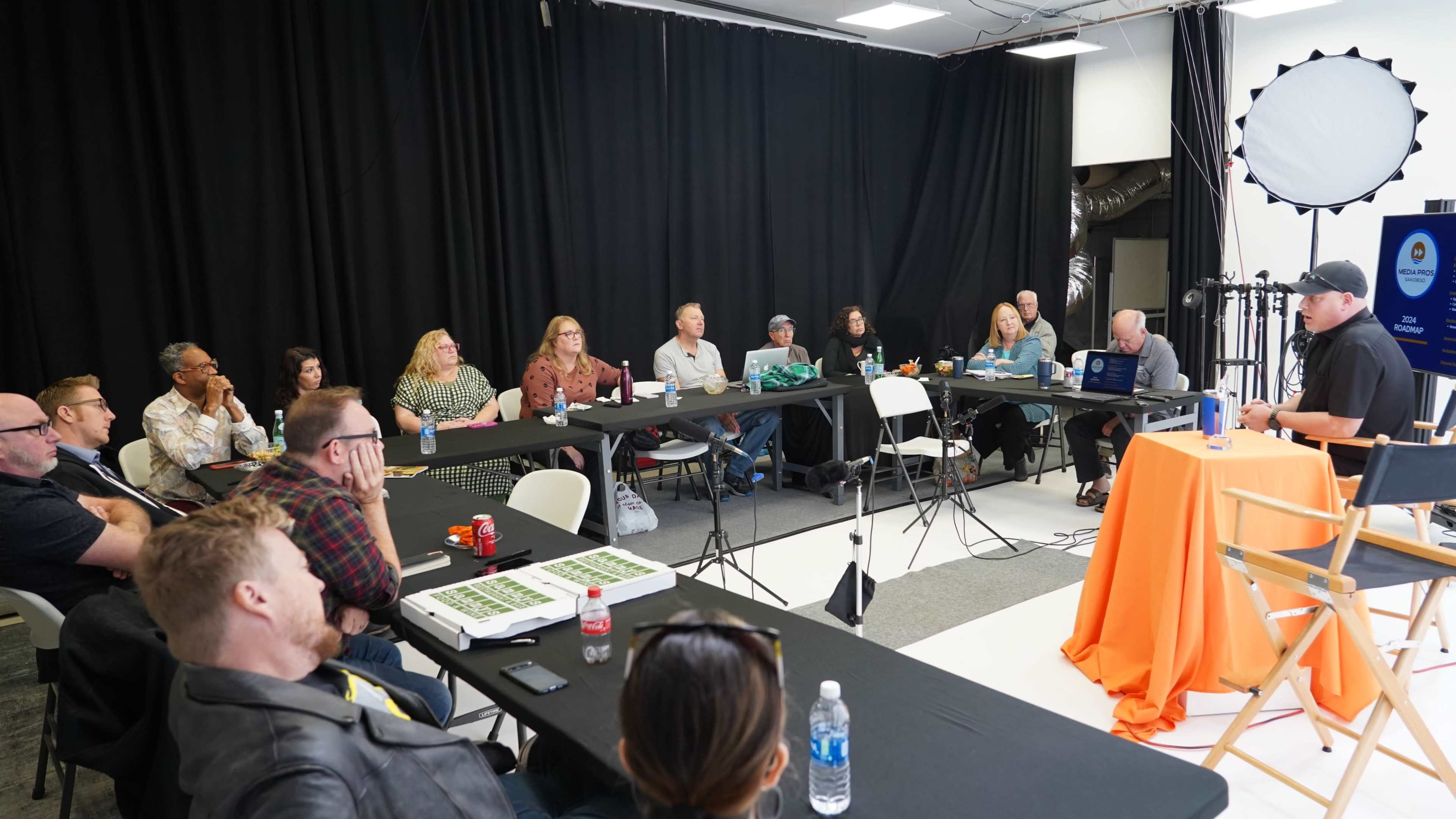 A group of people attends a meeting or workshop in a room with black curtains, sitting around tables equipped with laptops and materials.