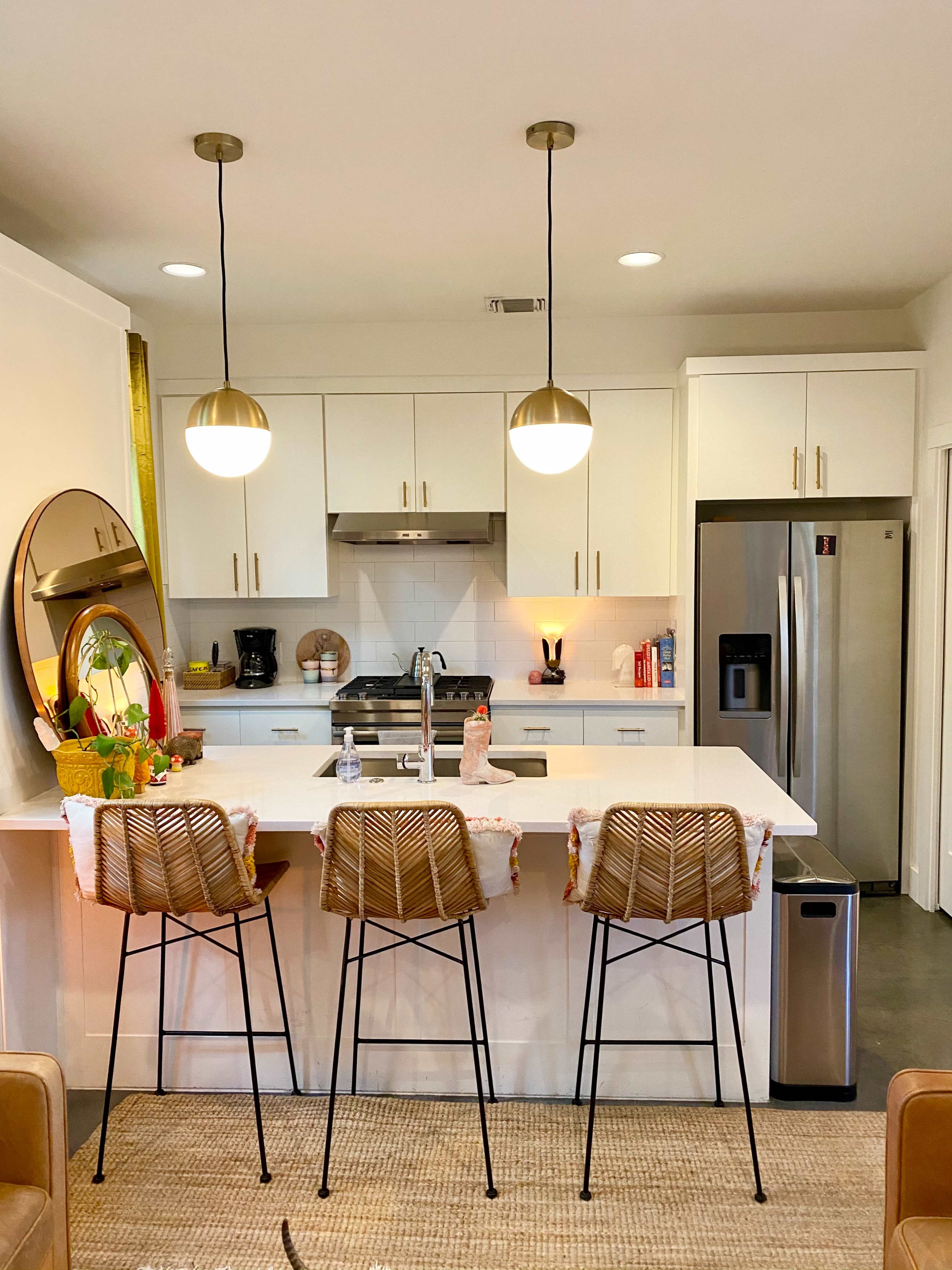 The image shows a modern kitchen with three bar stools at a breakfast counter, a stainless steel refrigerator, and overhead pendant lights.