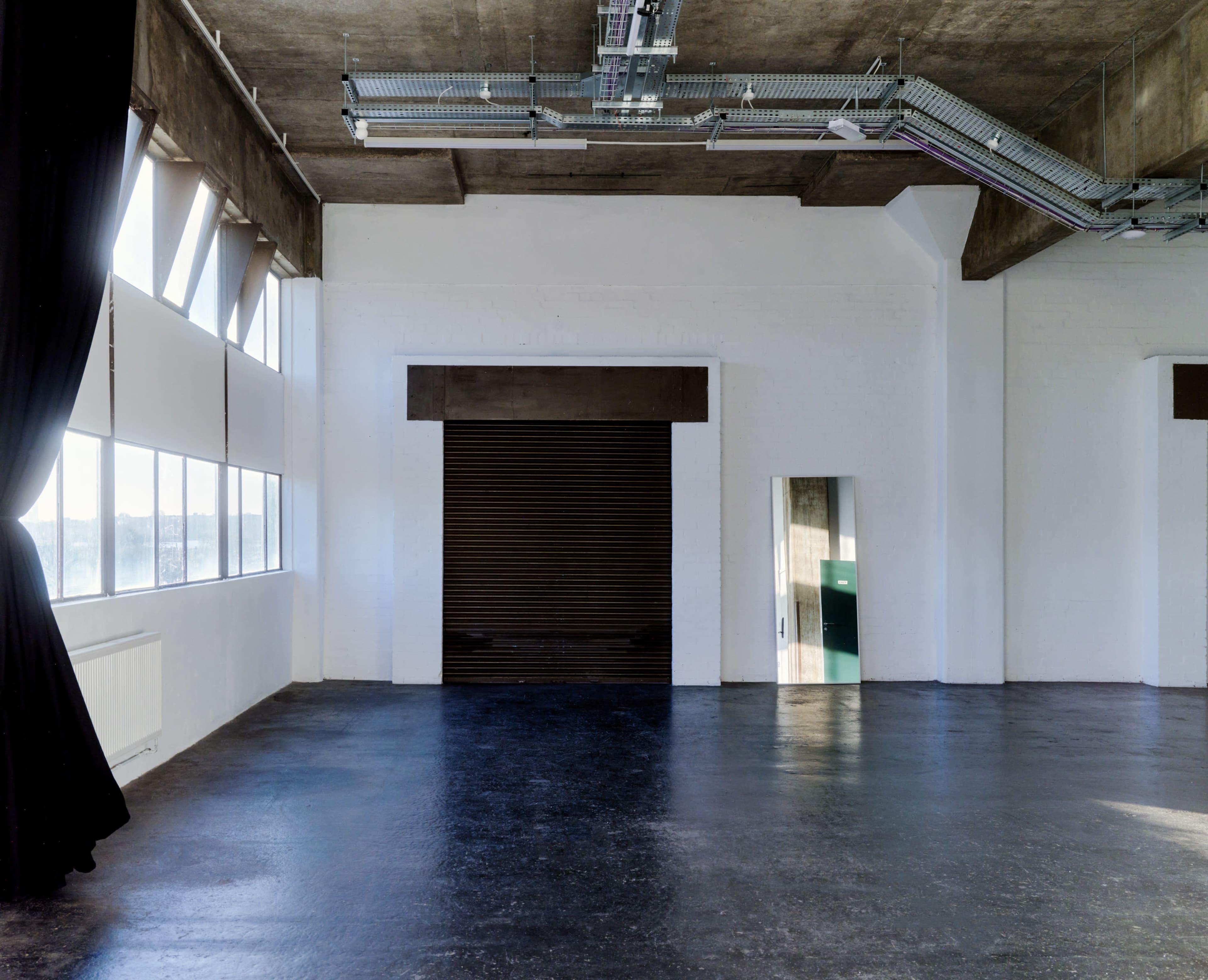 An empty industrial space with large windows, a roll-up door, and a mirror on the right wall.