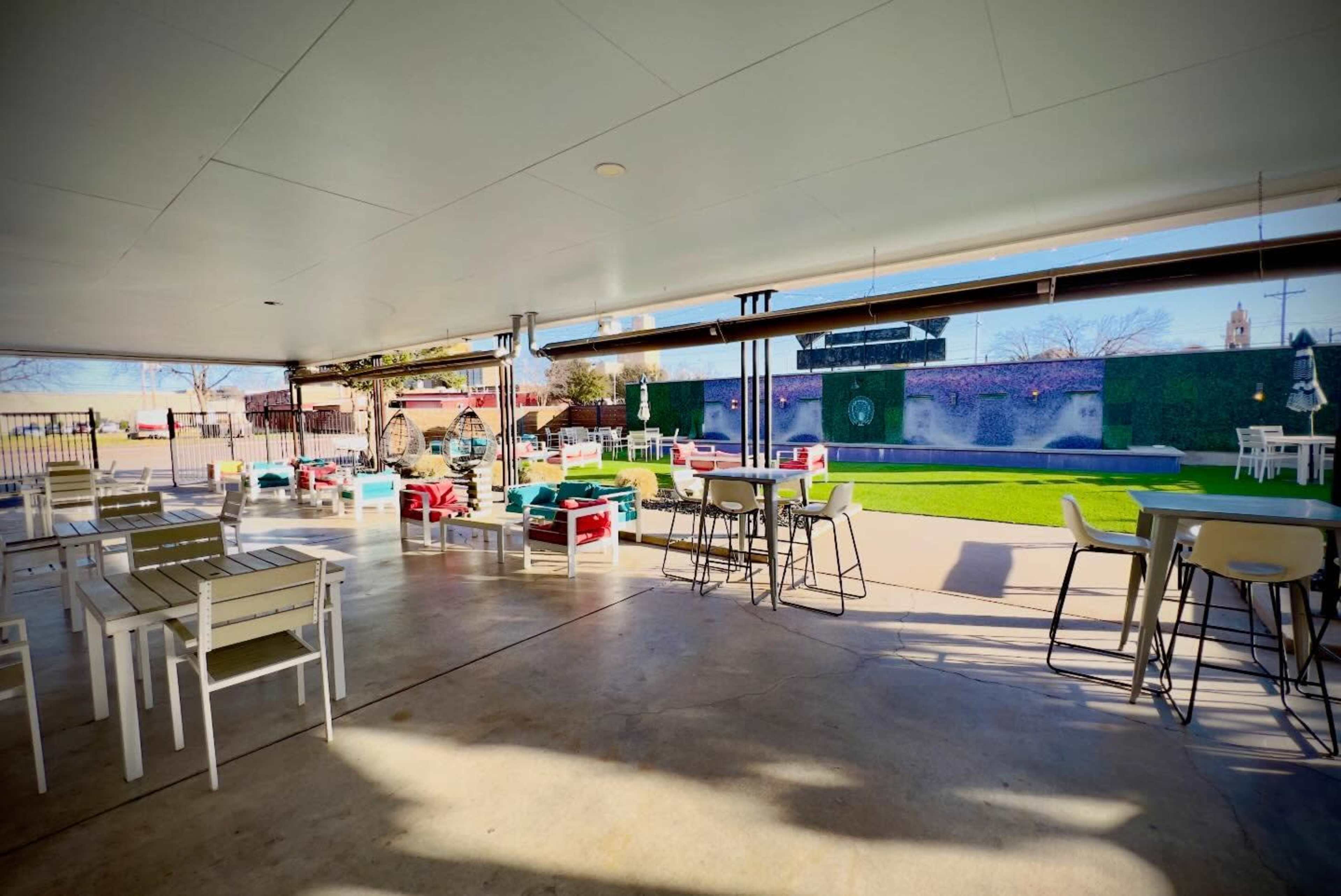 The image shows an outdoor seating area with tables and chairs under a shaded structure, overlooking a green lawn and a vibrant wall.