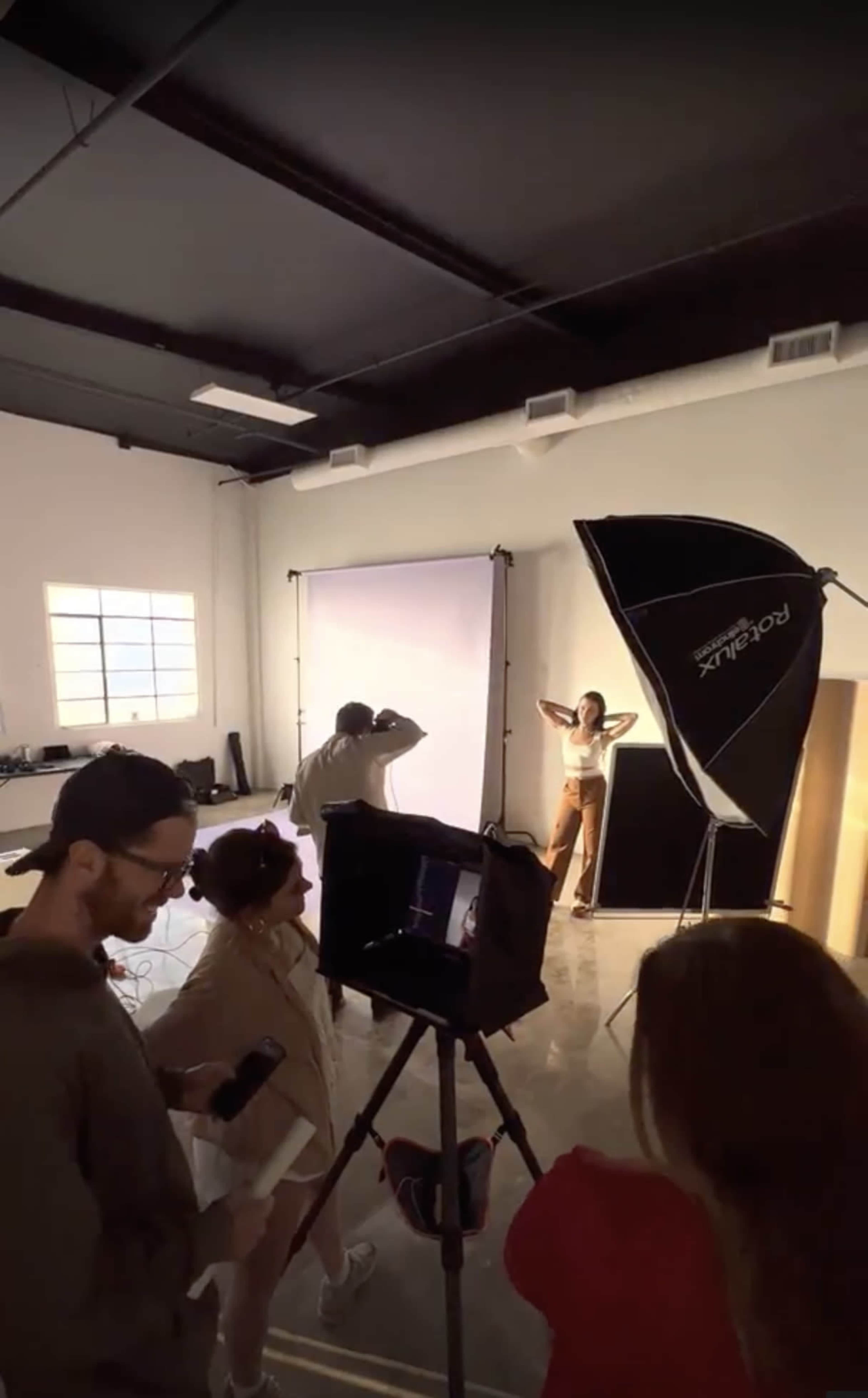 A group of people works in a photography studio, with a model posing under bright lights while photographers and assistants adjust equipment.