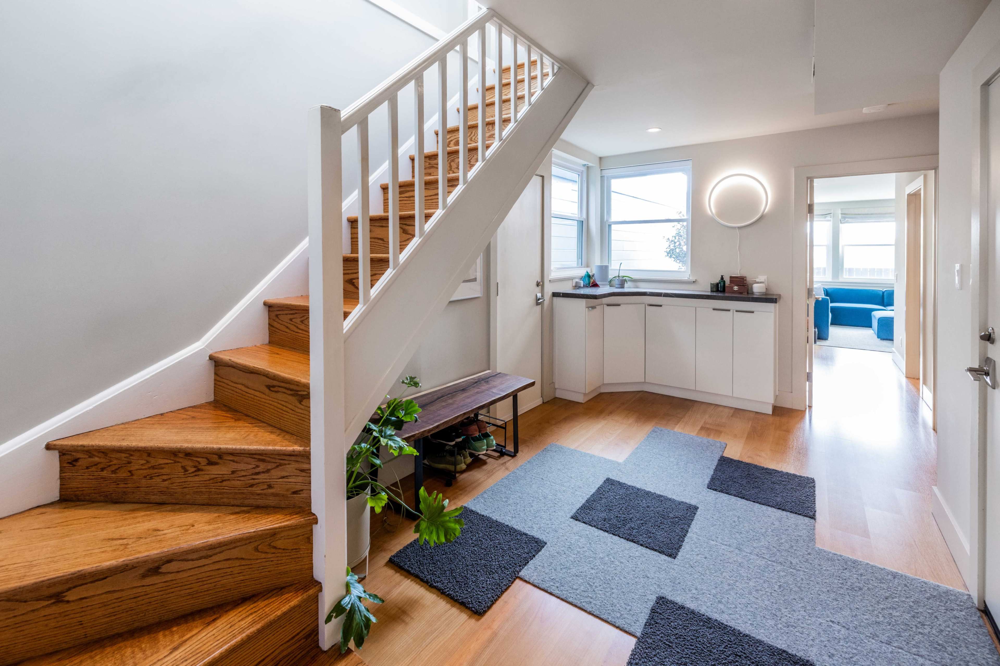 A bright entryway featuring a staircase on the left, a small kitchenette in the background, and a cozy seating area visible through an open doorway.