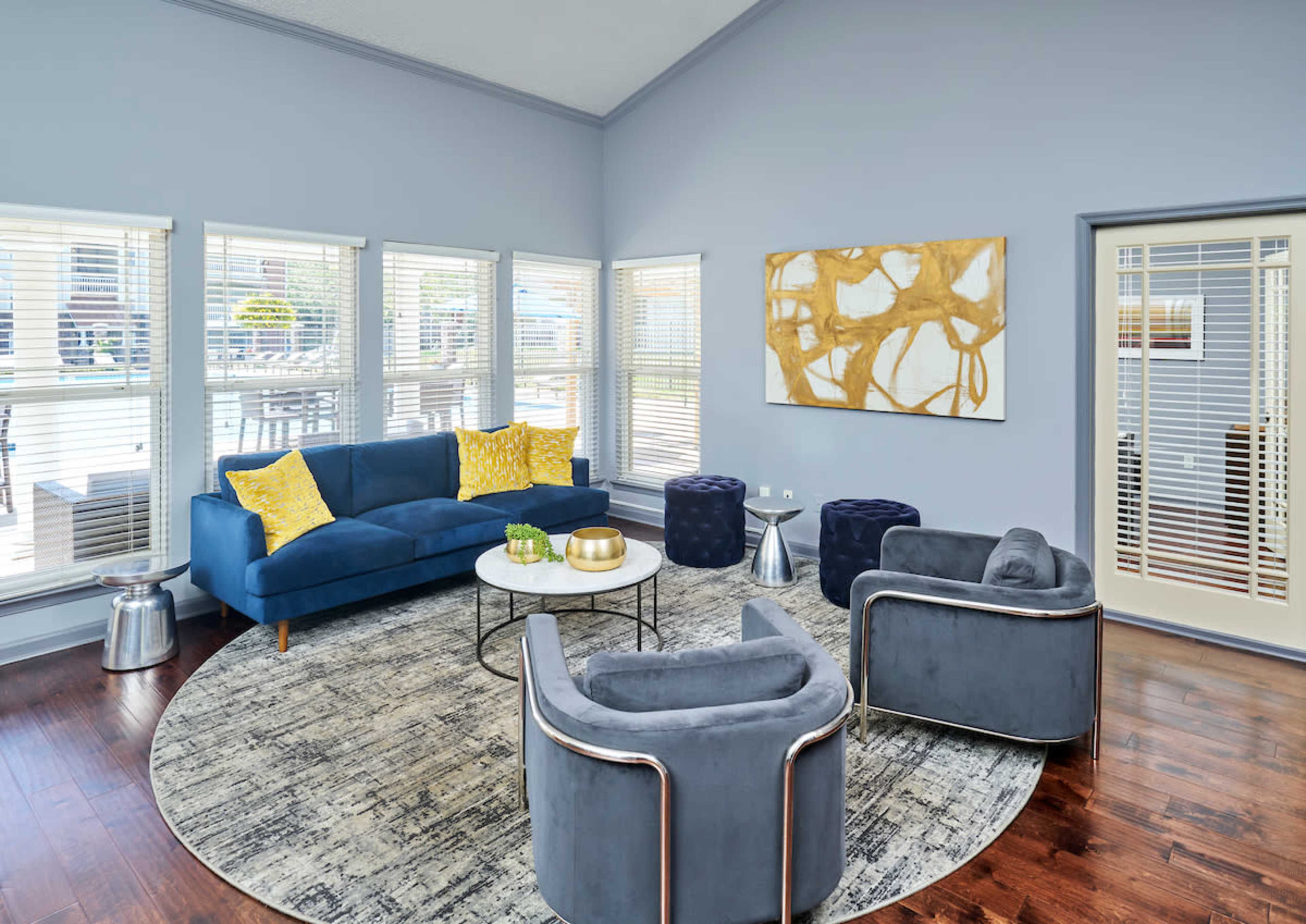 A modern living room features a blue sofa, two round chairs, a circular rug, and large windows with blinds.
