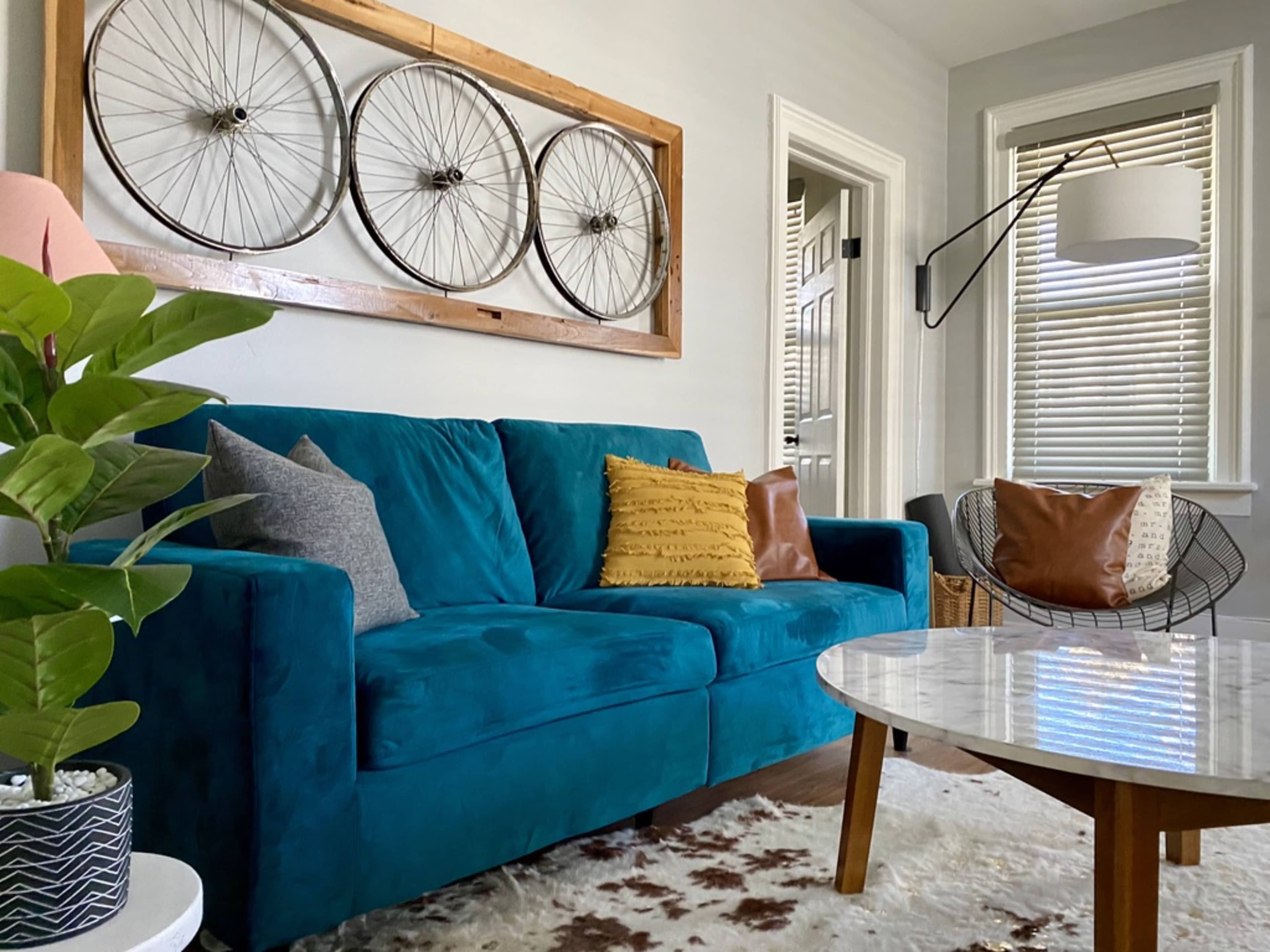 A vibrant teal couch is positioned in a living room adorned with a decorative arrangement of bicycle wheels on the wall, accompanied by a round marble coffee table and various decorative accents.
