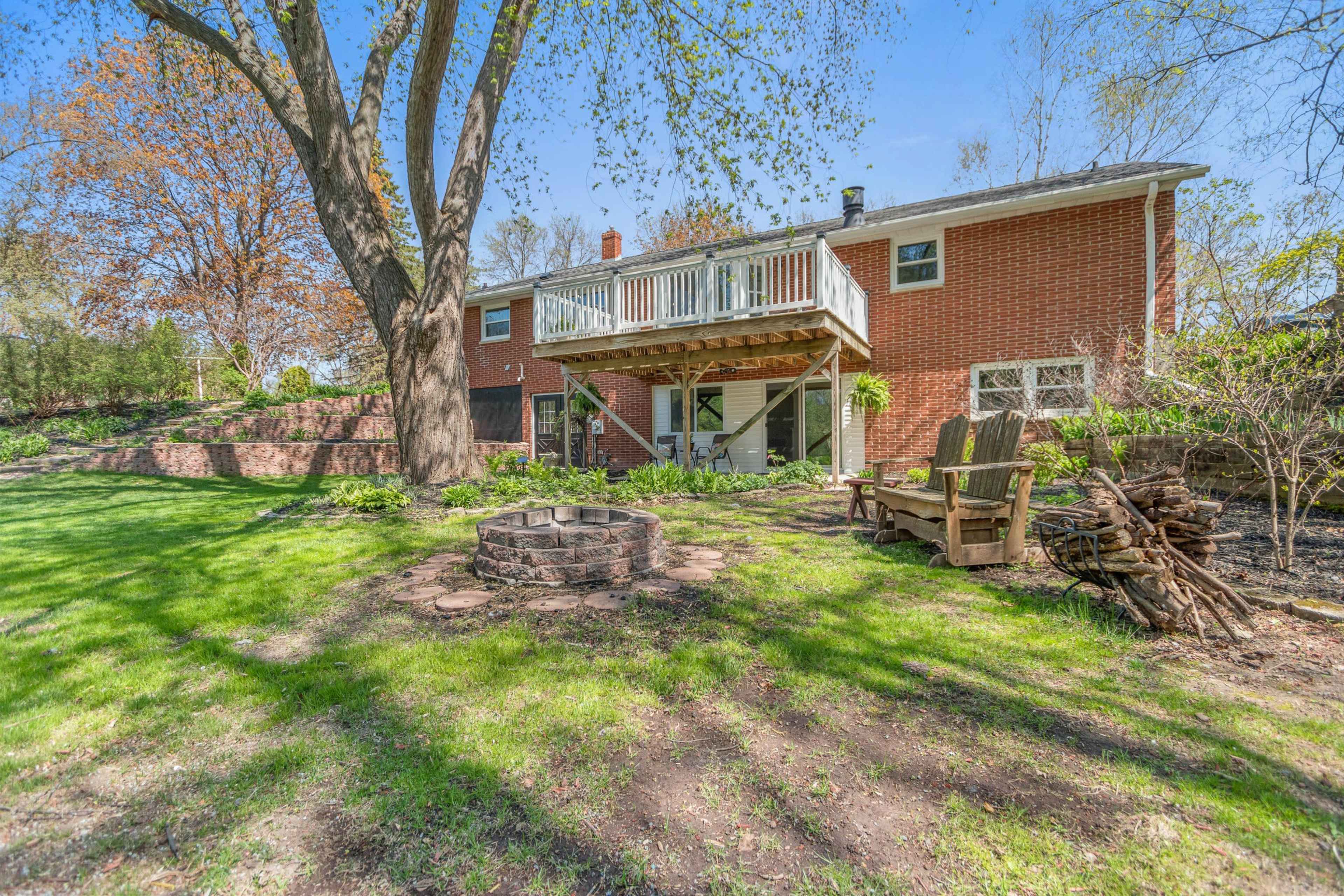 A brick house with a balcony overlooking a green yard that features a fire pit and stacked firewood.