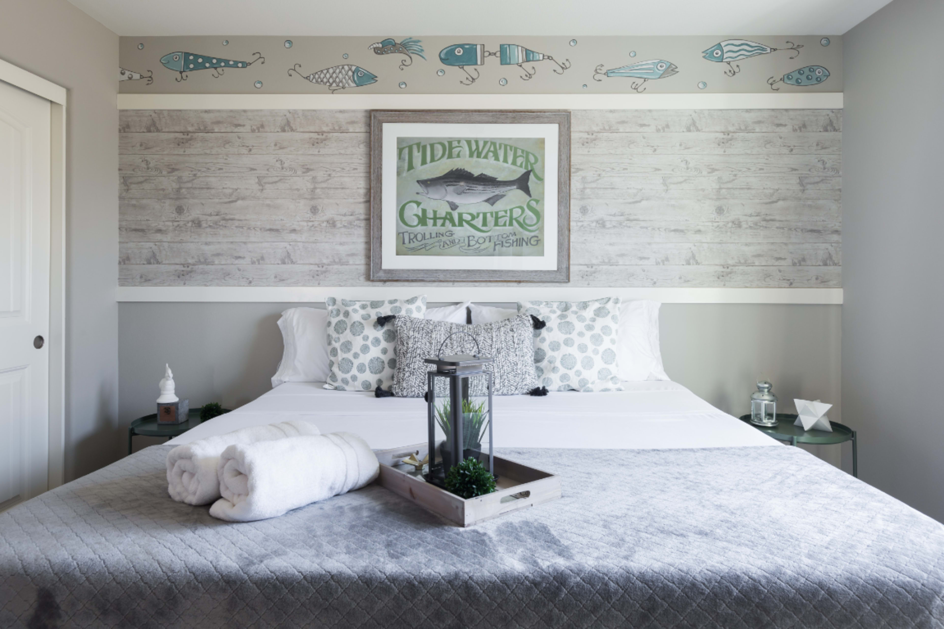 A neatly arranged bedroom featuring a bed with decorative pillows, a tray with towels and plants, and a fishing-themed wall art above the headboard.