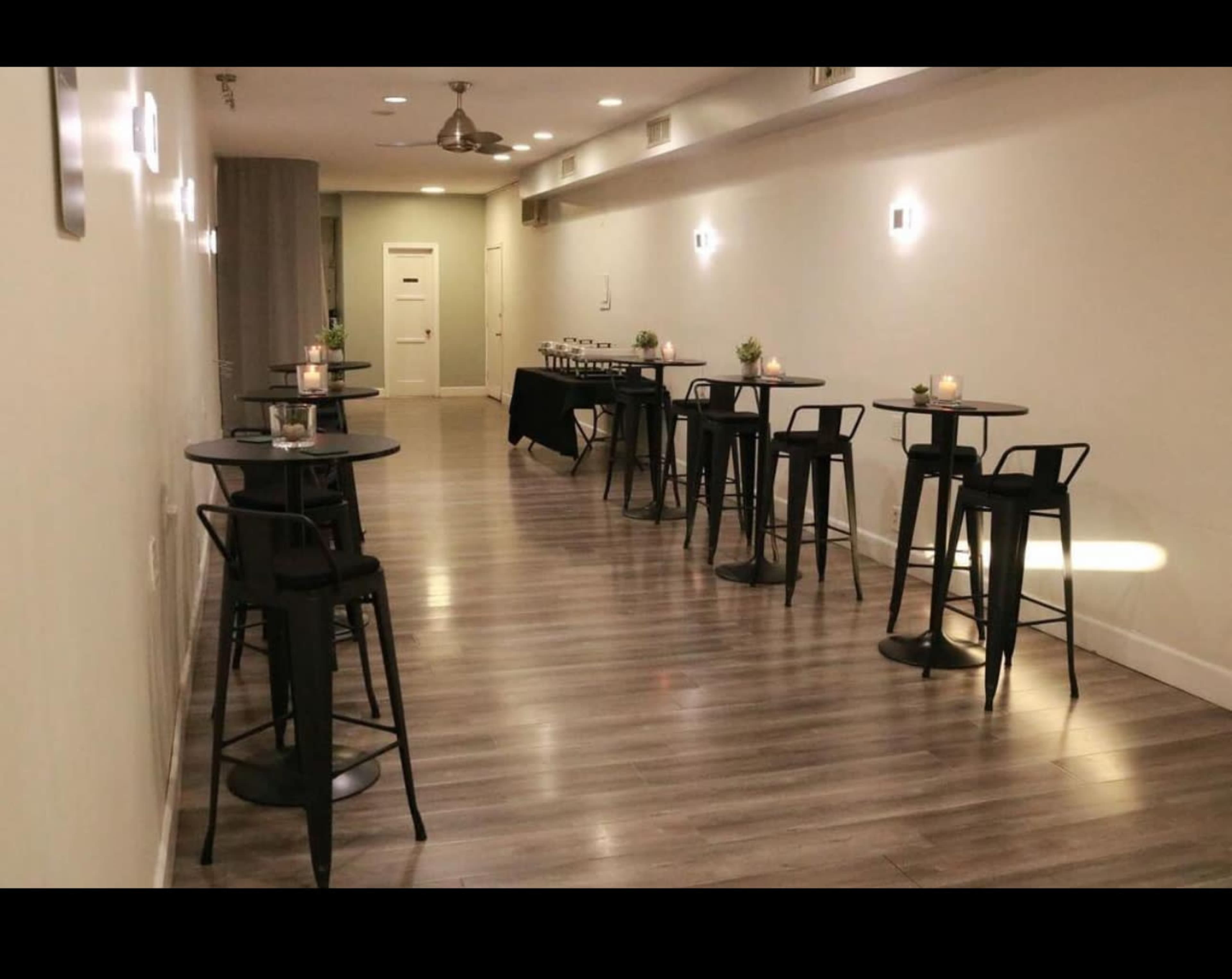 The image shows a modern, well-lit event space with several tall, round tables and chairs arranged neatly along a long wall.