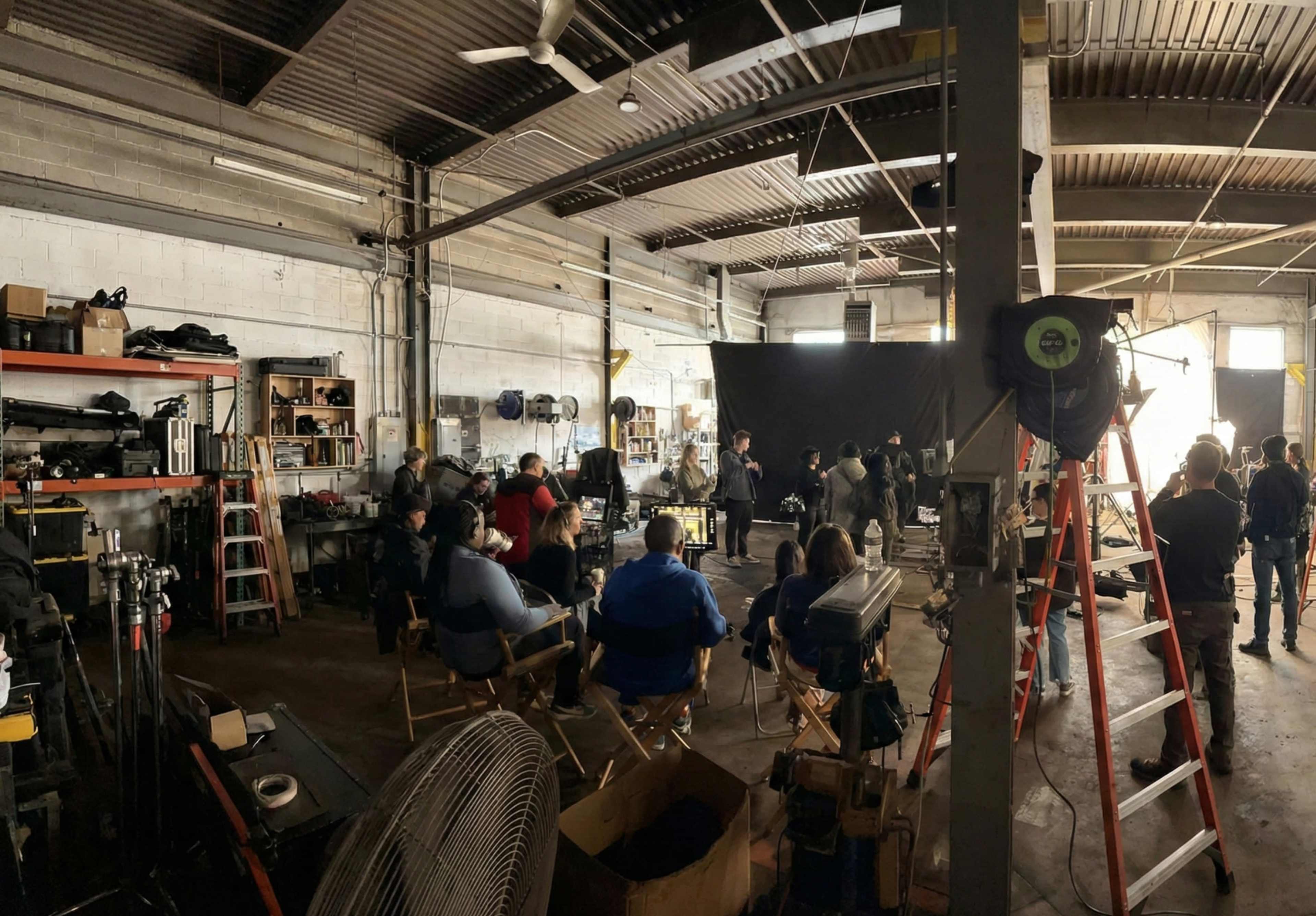A film crew and audience are gathered in a spacious warehouse with various equipment and shelves visible in the background.
