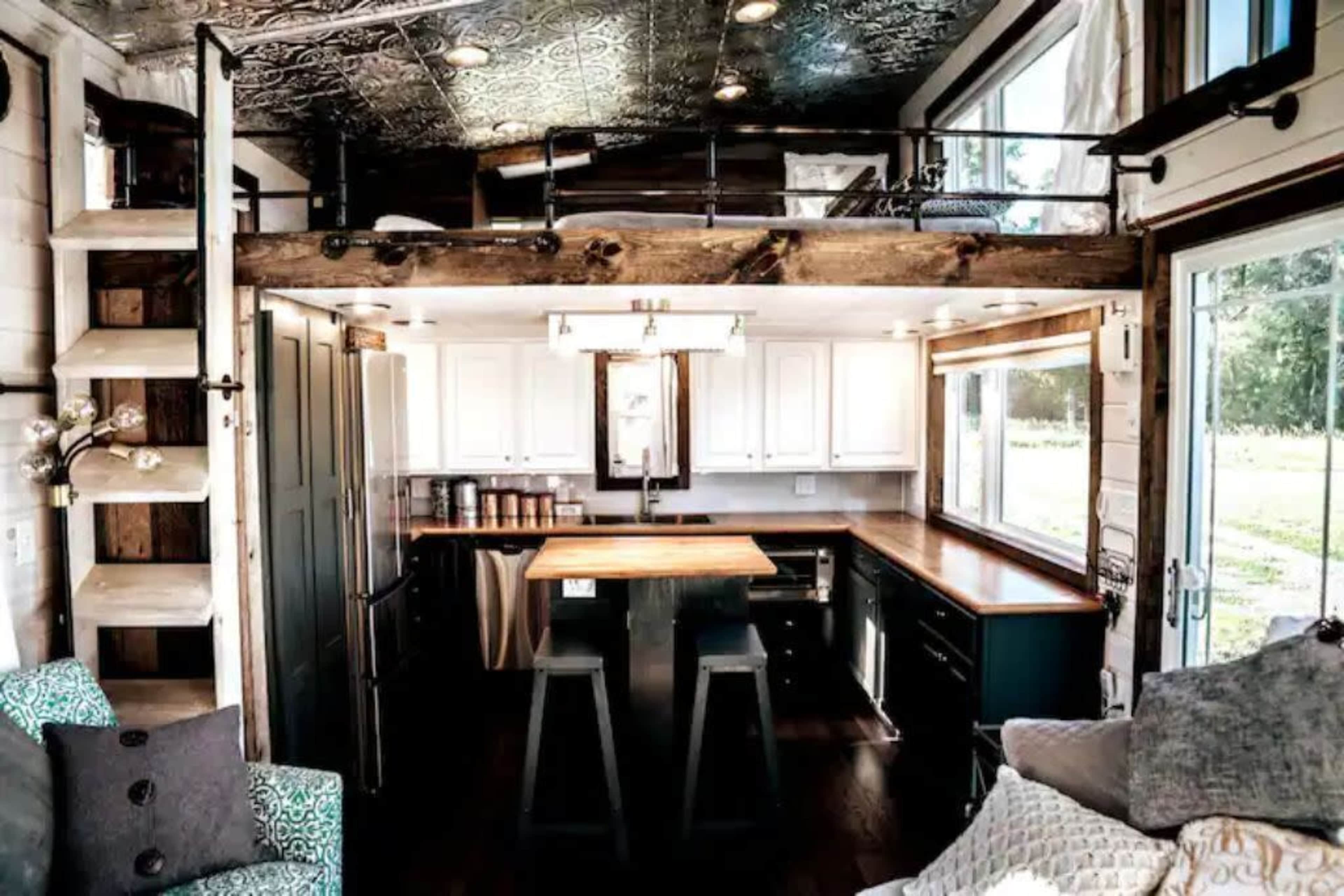 A modern tiny house interior with a lofted sleeping area, a kitchen island, and large windows that provide natural light.