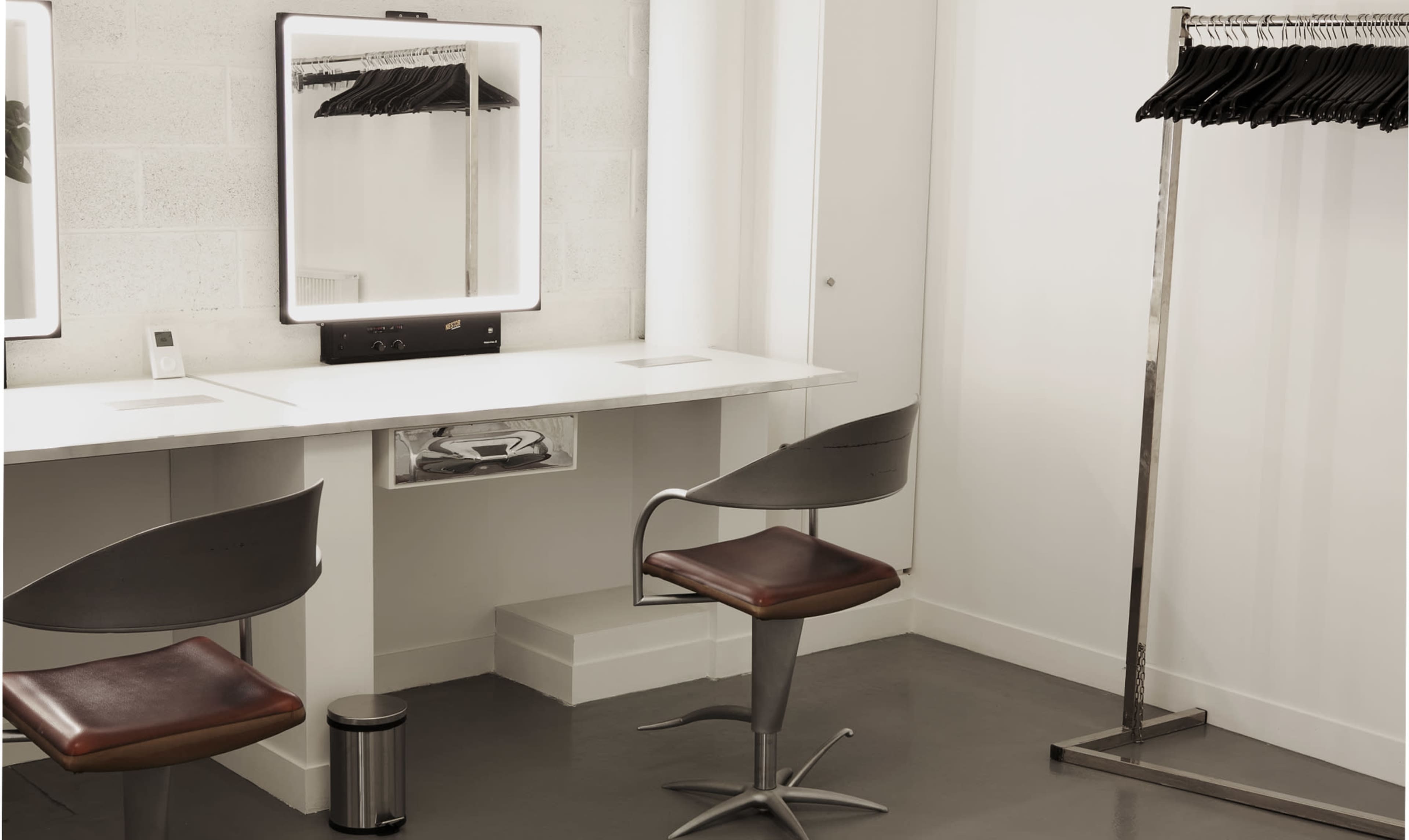 The image shows a minimalist dressing room with a two-seat vanity, a mirror with a light, and a clothing rack beside it.