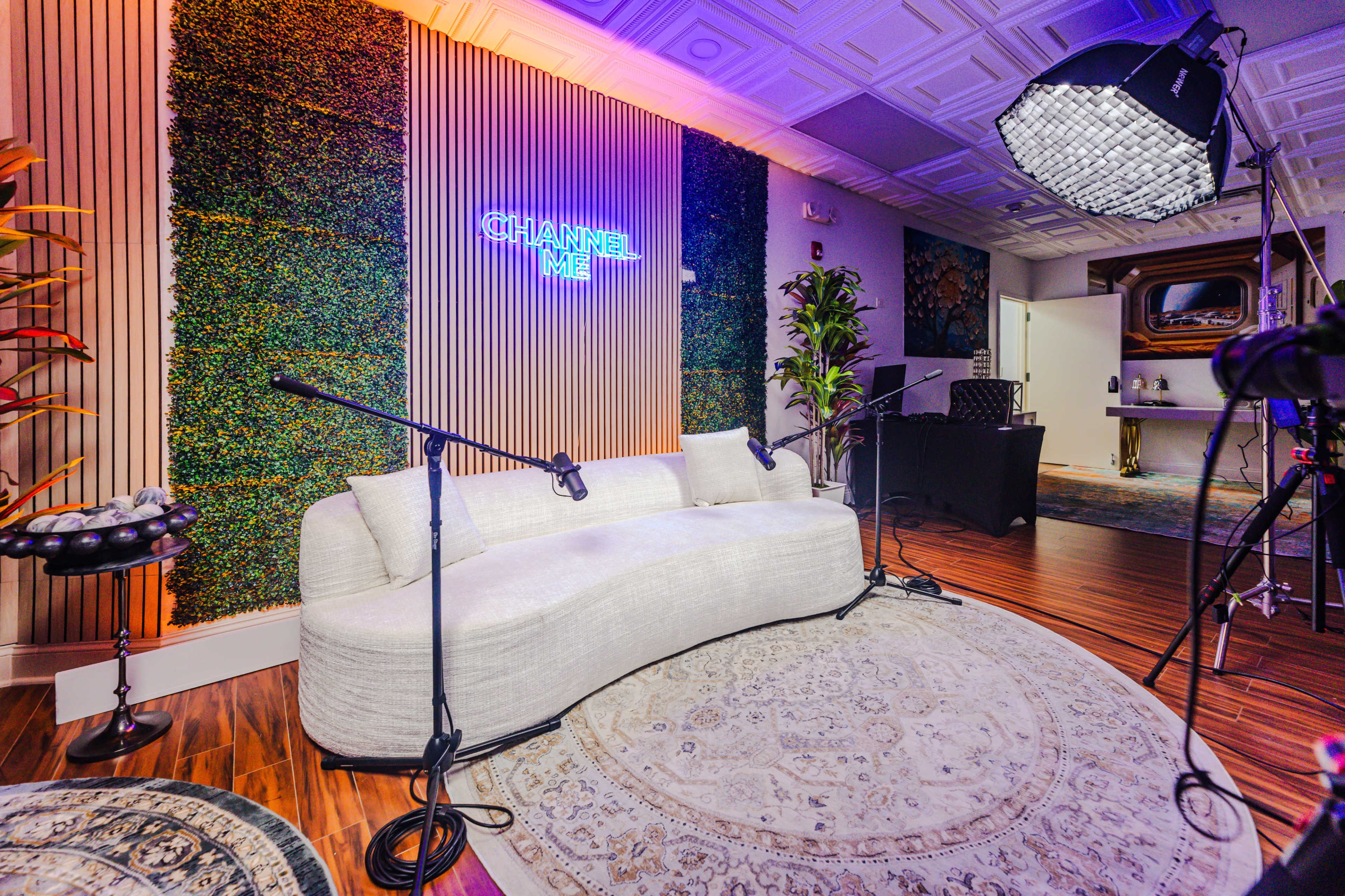The image shows a modern recording studio with a curved sofa, green wall decor, and a neon sign that reads "CHANNEL ME," alongside microphones and studio equipment set up for use.