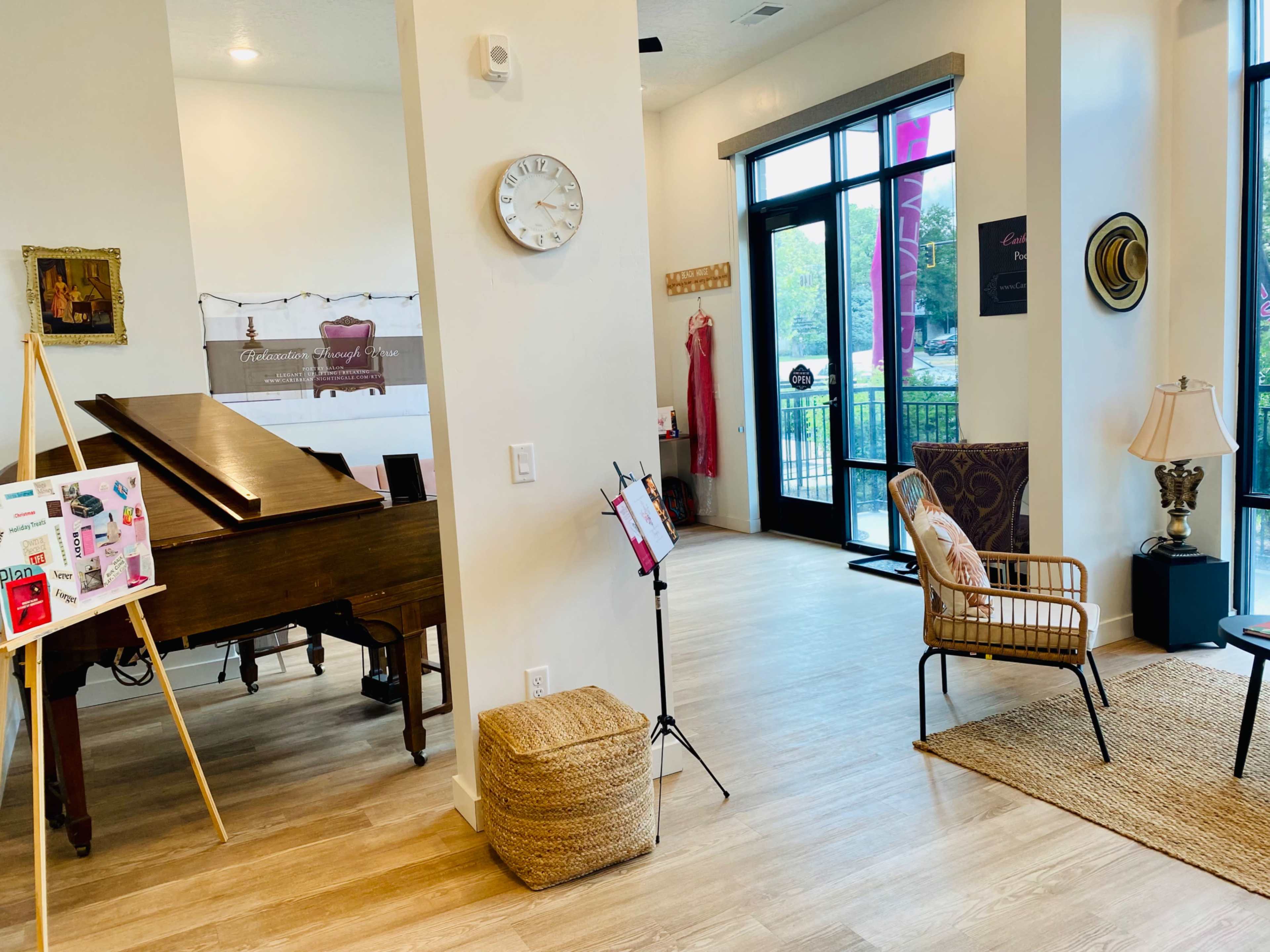 An indoor space featuring a grand piano, a wall clock, a wicker chair, and large windows with a view outside.