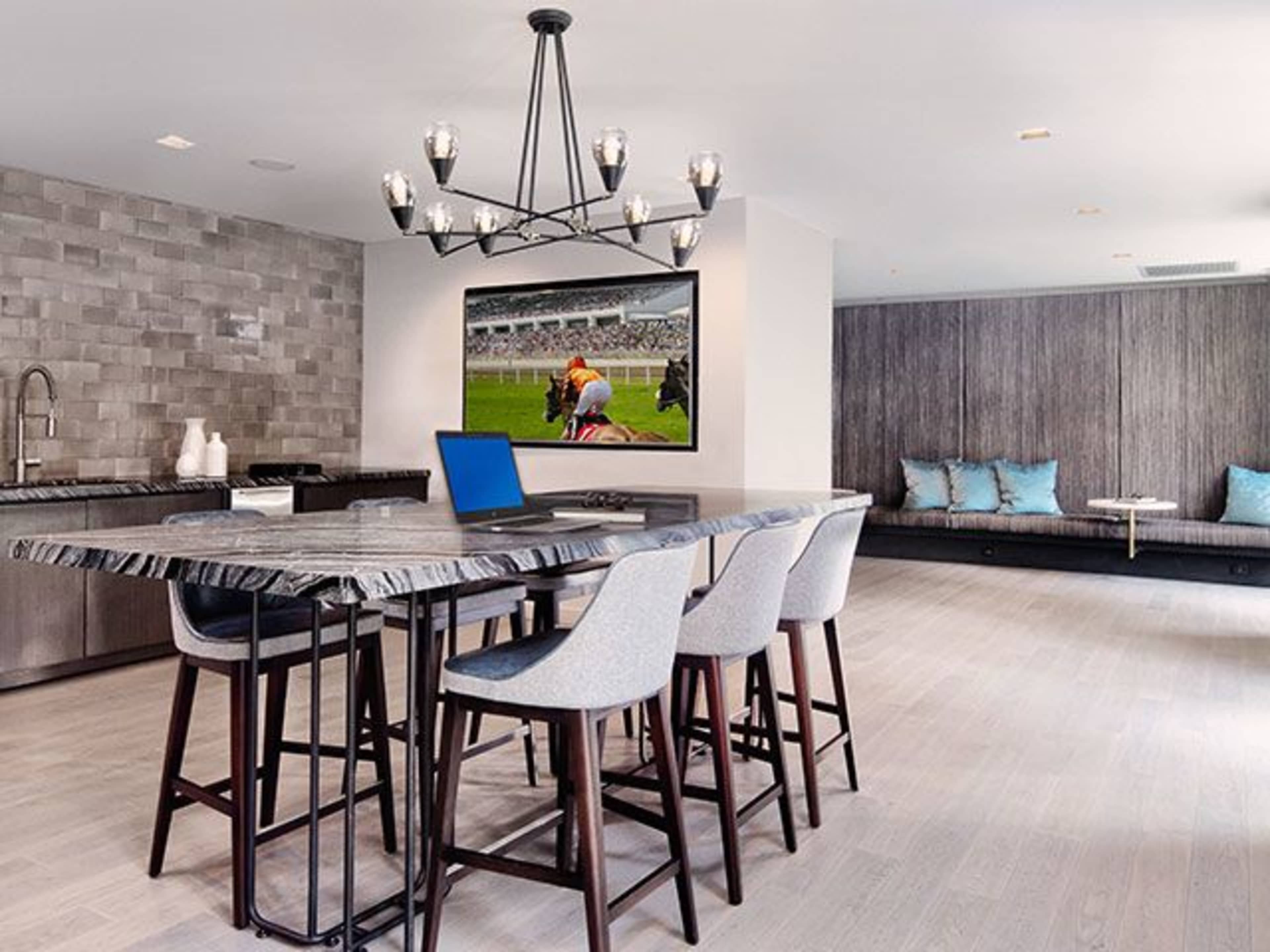 The image shows a modern kitchen or dining area with a large island, several high stools, a wall-mounted television displaying a sports event, and a cozy seating area in the background.