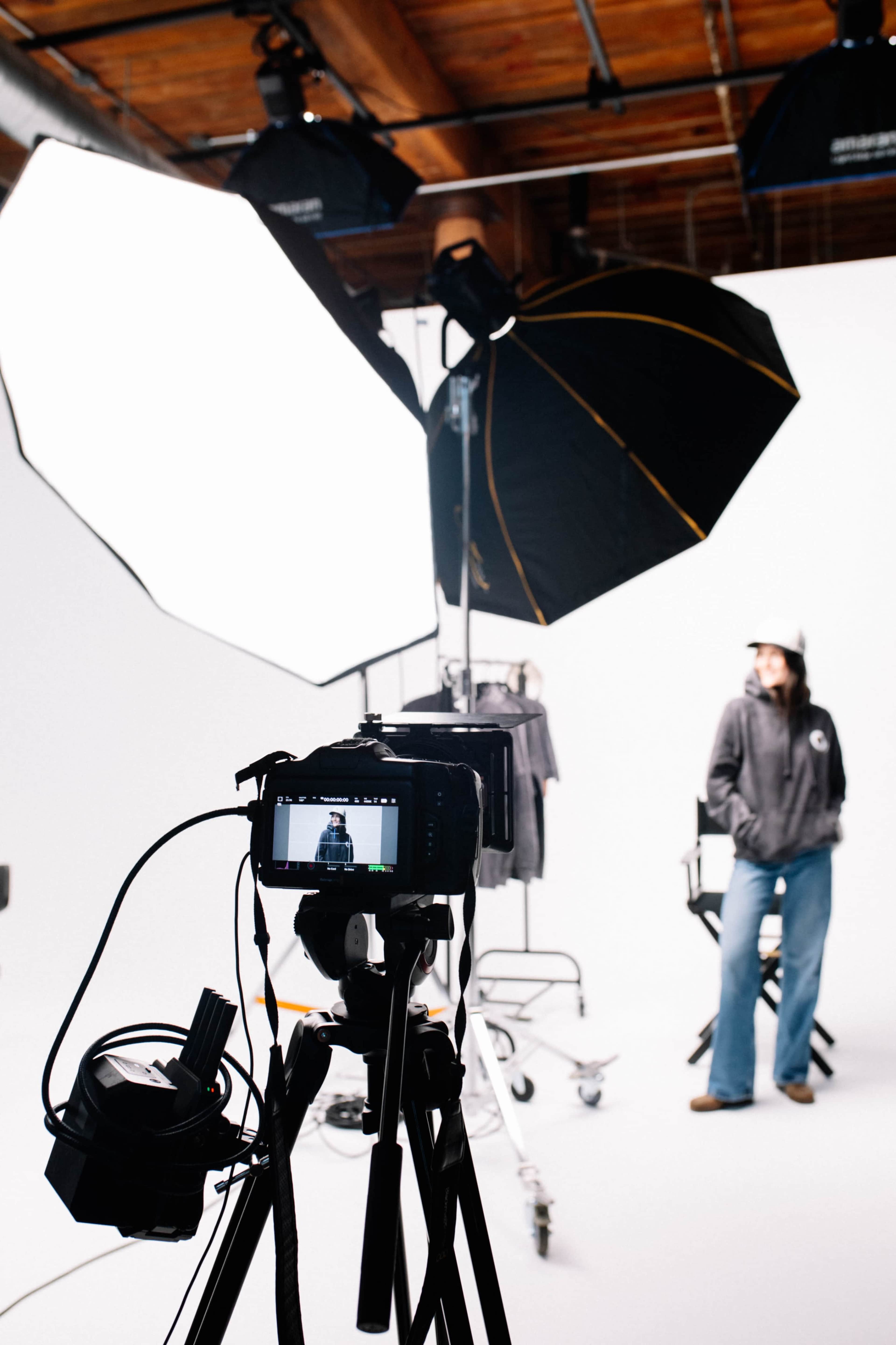 A camera on a tripod is positioned in front of a white backdrop, capturing a person sitting on a director's chair under studio lighting.