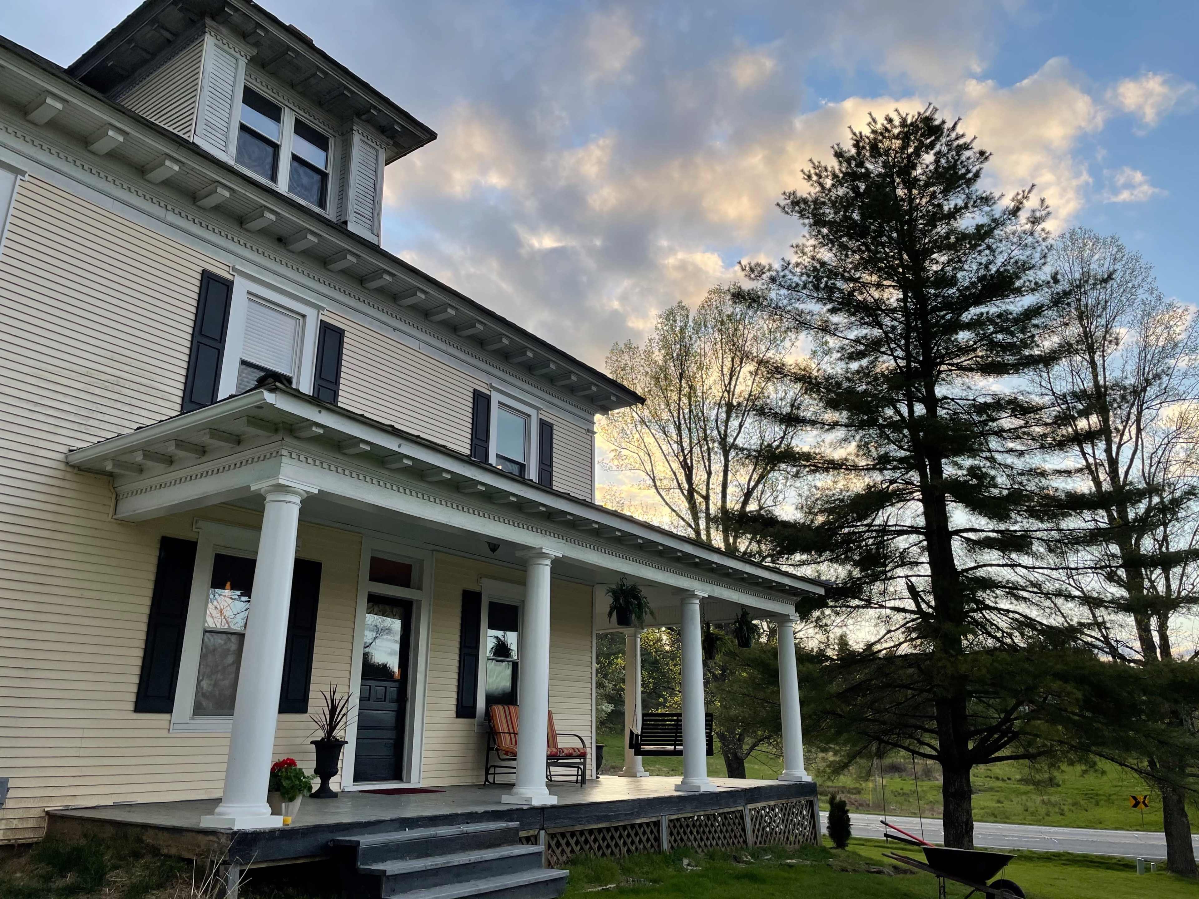 A large, two-story house with white siding and a covered front porch is positioned next to a tall evergreen tree under a partly cloudy sky.