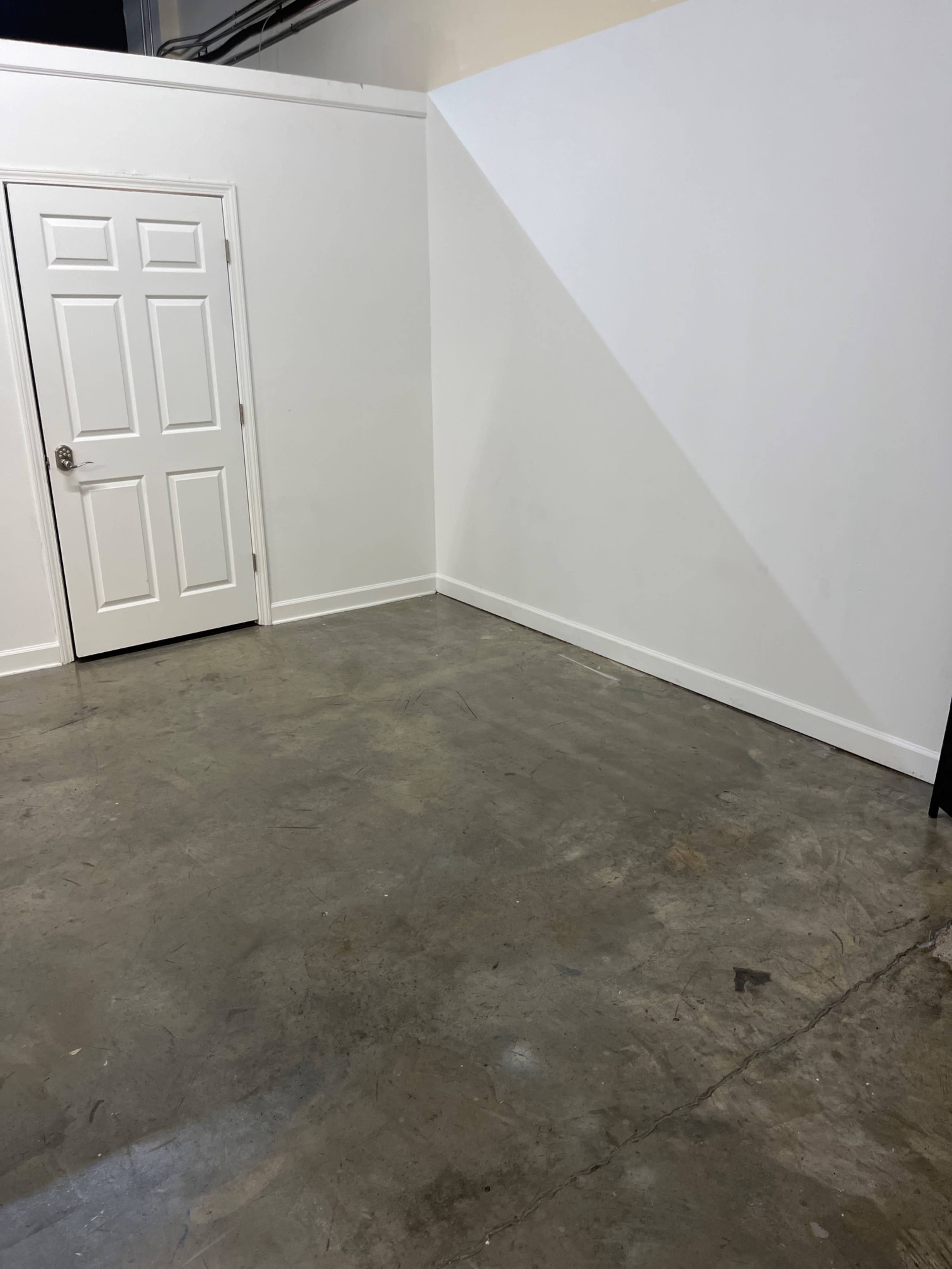 The image shows a vacant room with a white wall, a door on the left, and a concrete floor.