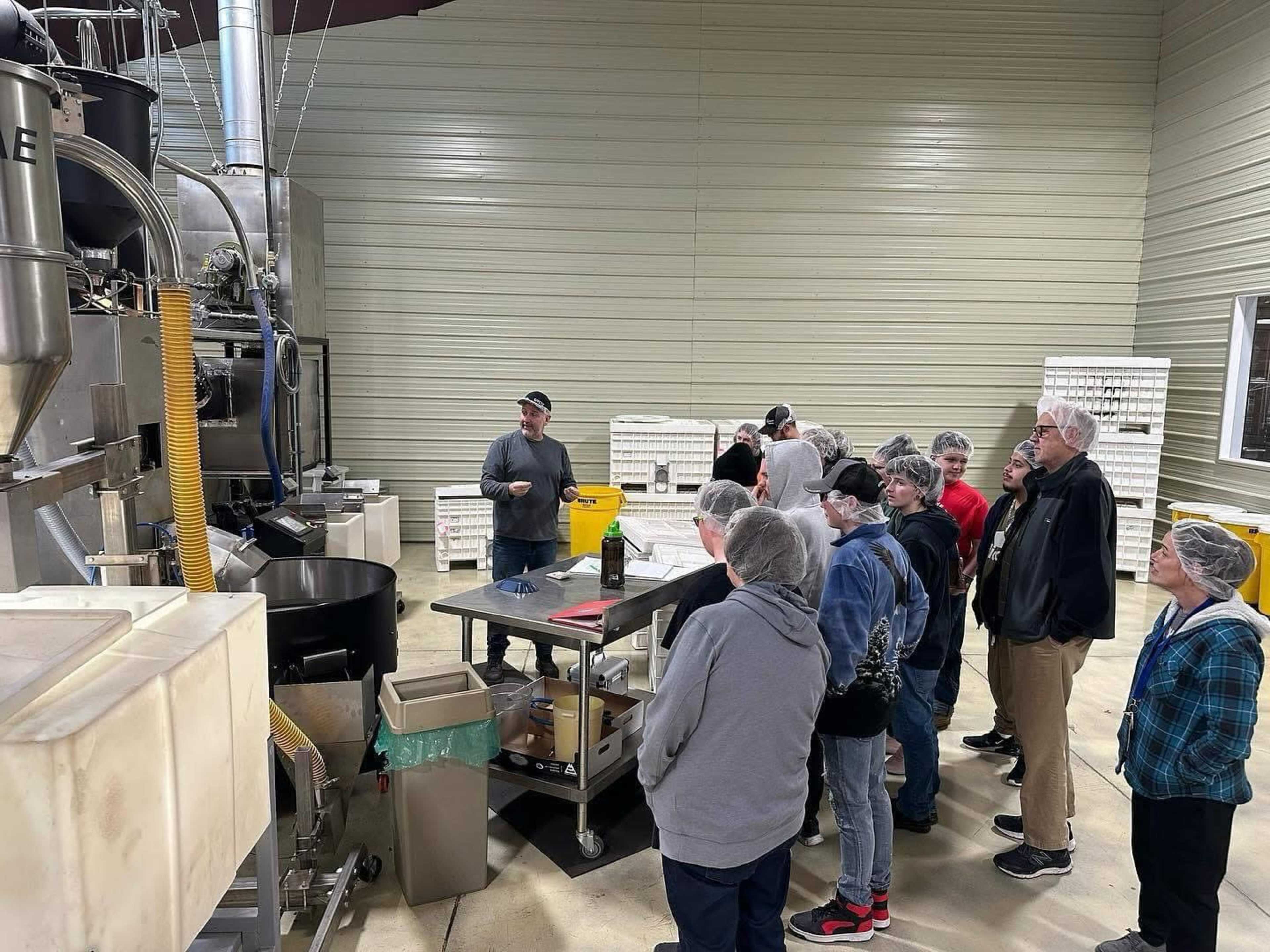 A group of people in hairnets watches a demonstration at a processing facility.