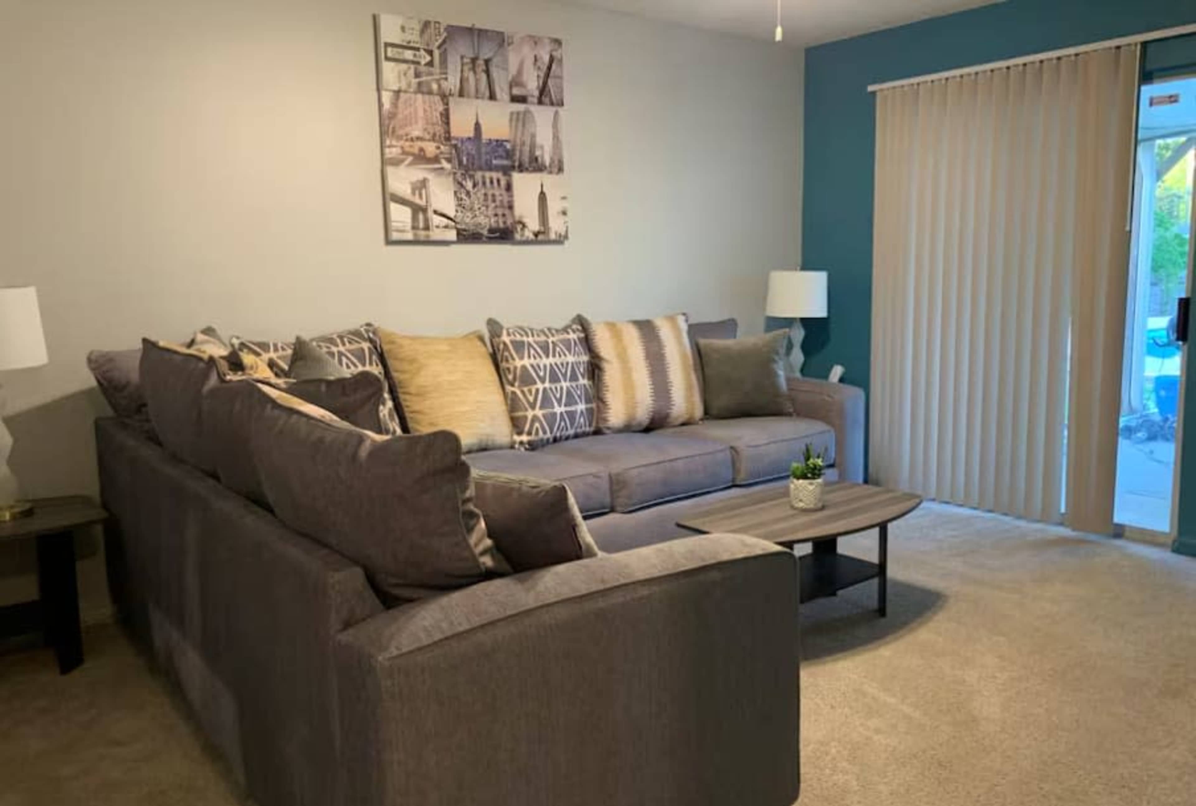 The image shows a cozy living room with a large gray sectional couch, decorative pillows, a small coffee table, and a sliding glass door with vertical blinds.