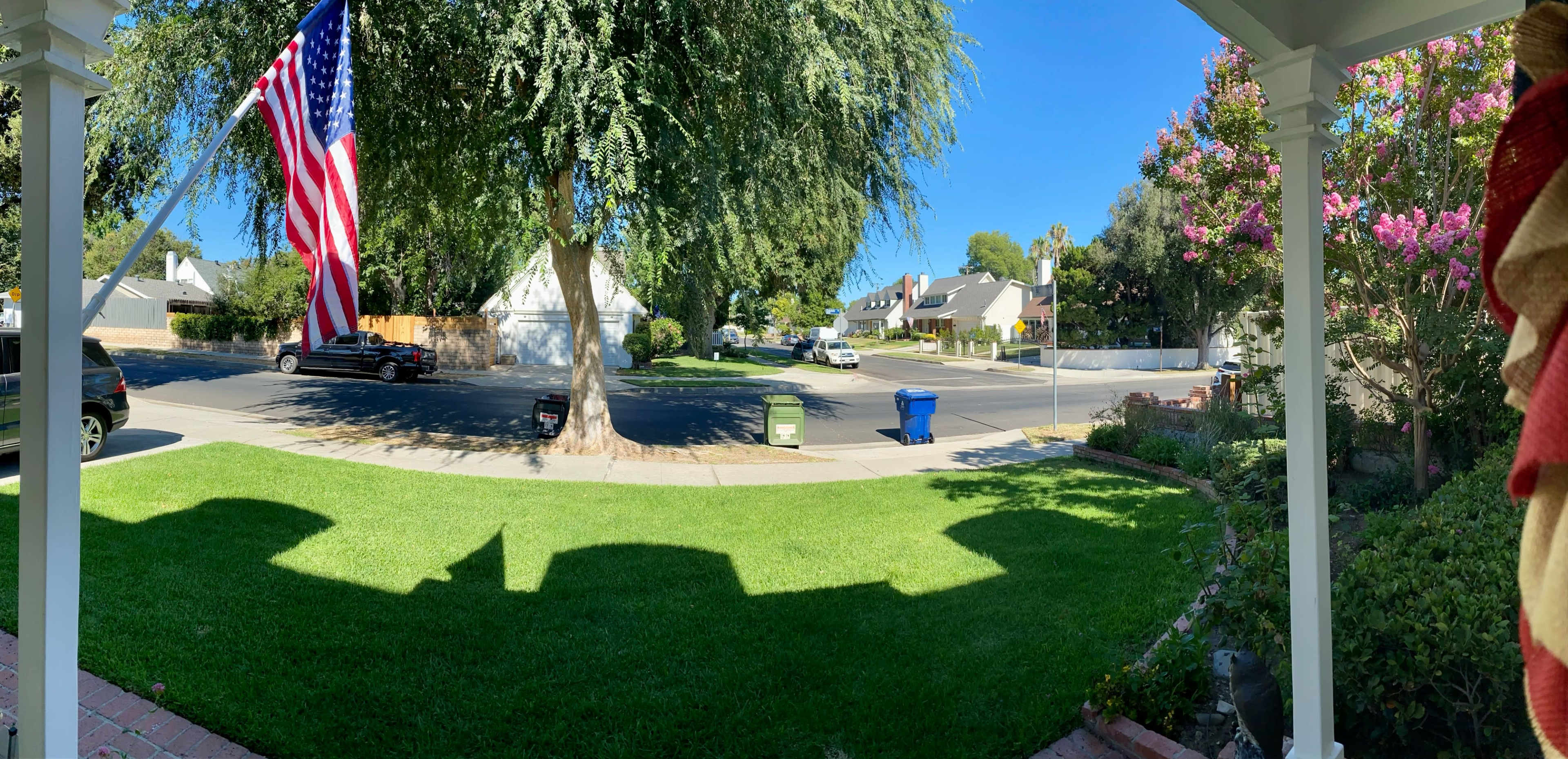 The image shows a residential street with houses, a large tree, and a visible American flag, all viewed from a porch with shadows cast on the lawn.