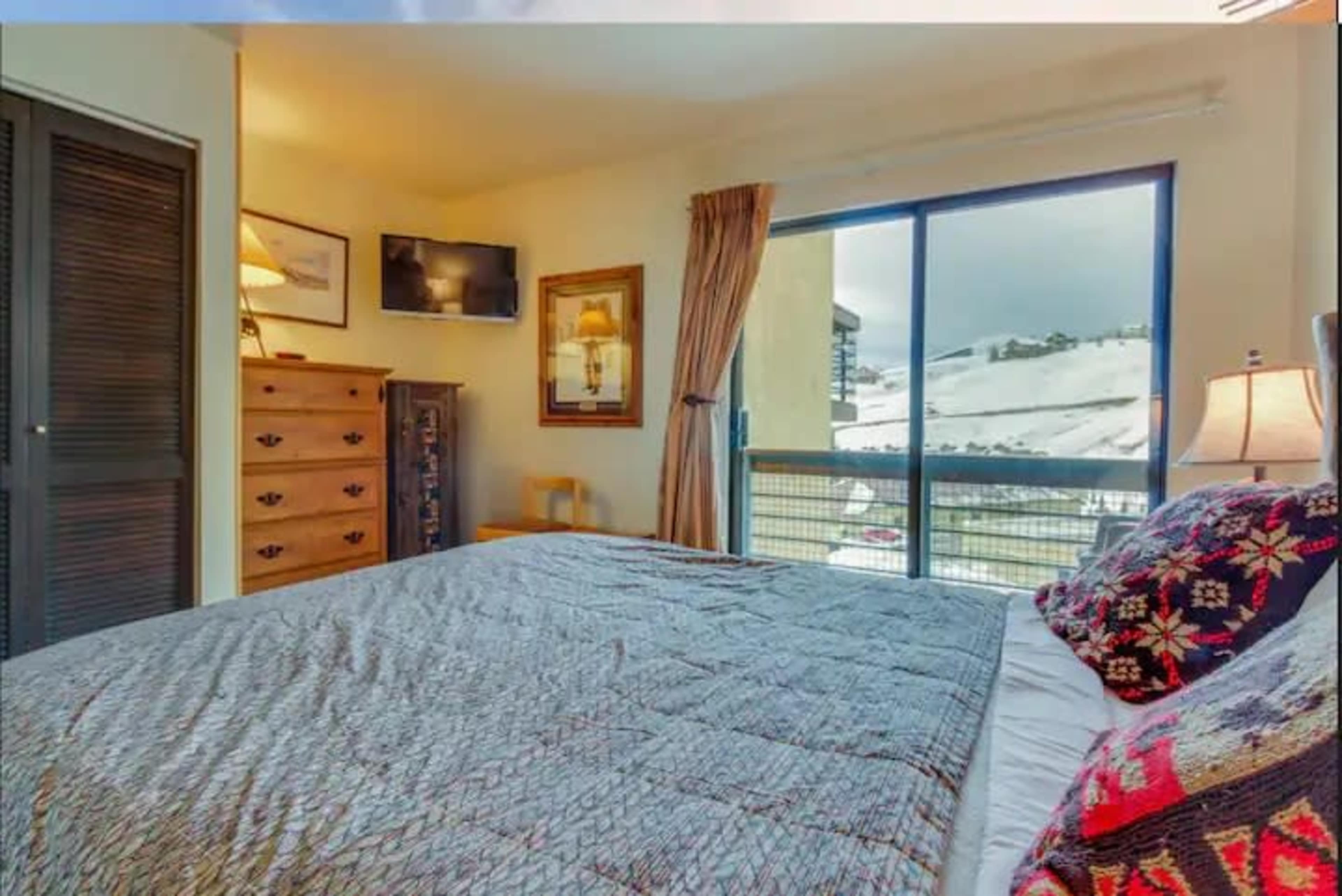 A bedroom featuring a large bed with a patterned duvet, a wooden dresser, a wall-mounted TV, and a window displaying a snowy landscape.