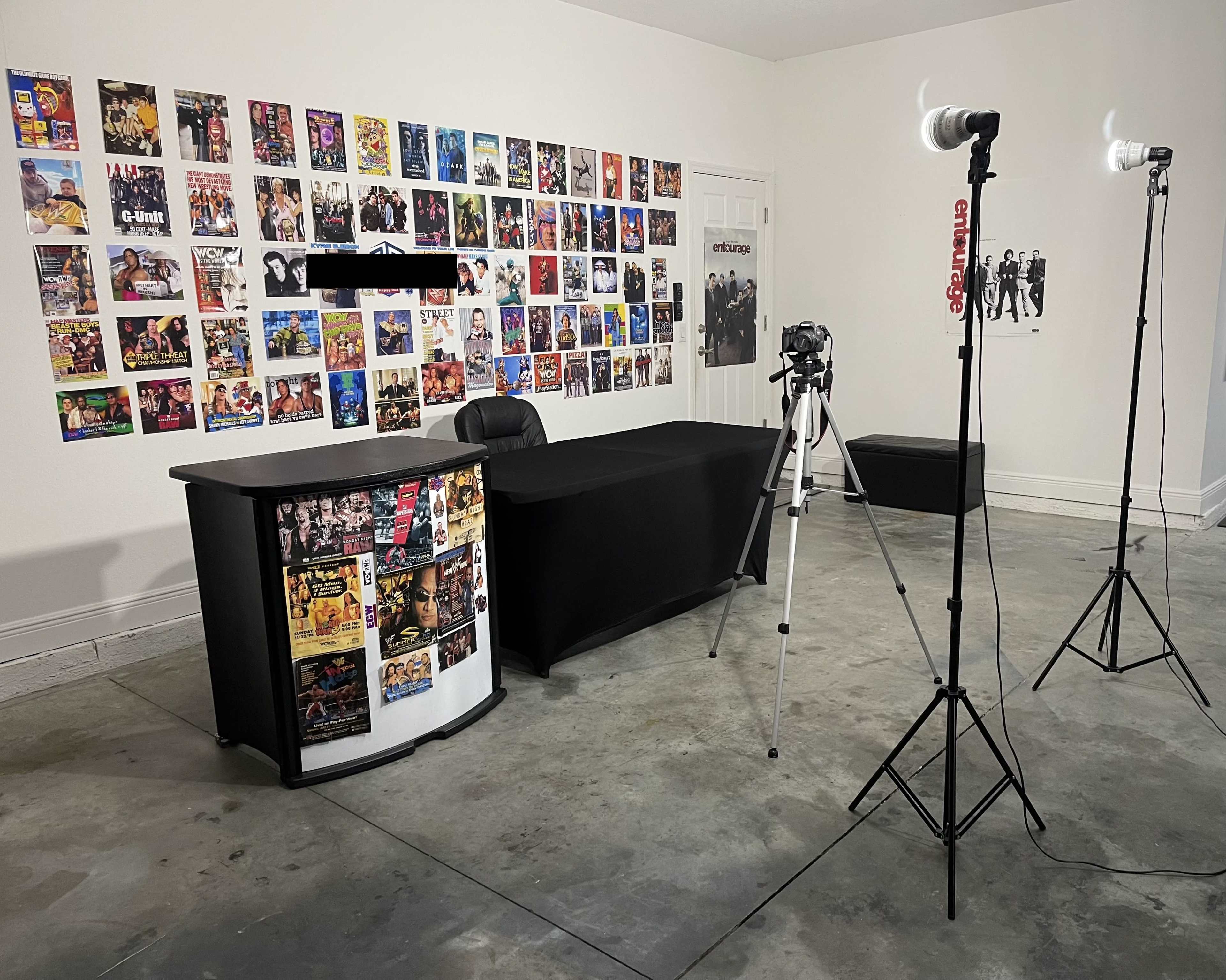 The image shows a setup for a photo or video shoot, featuring a desk with posters on the wall behind it and lighting equipment positioned nearby.
