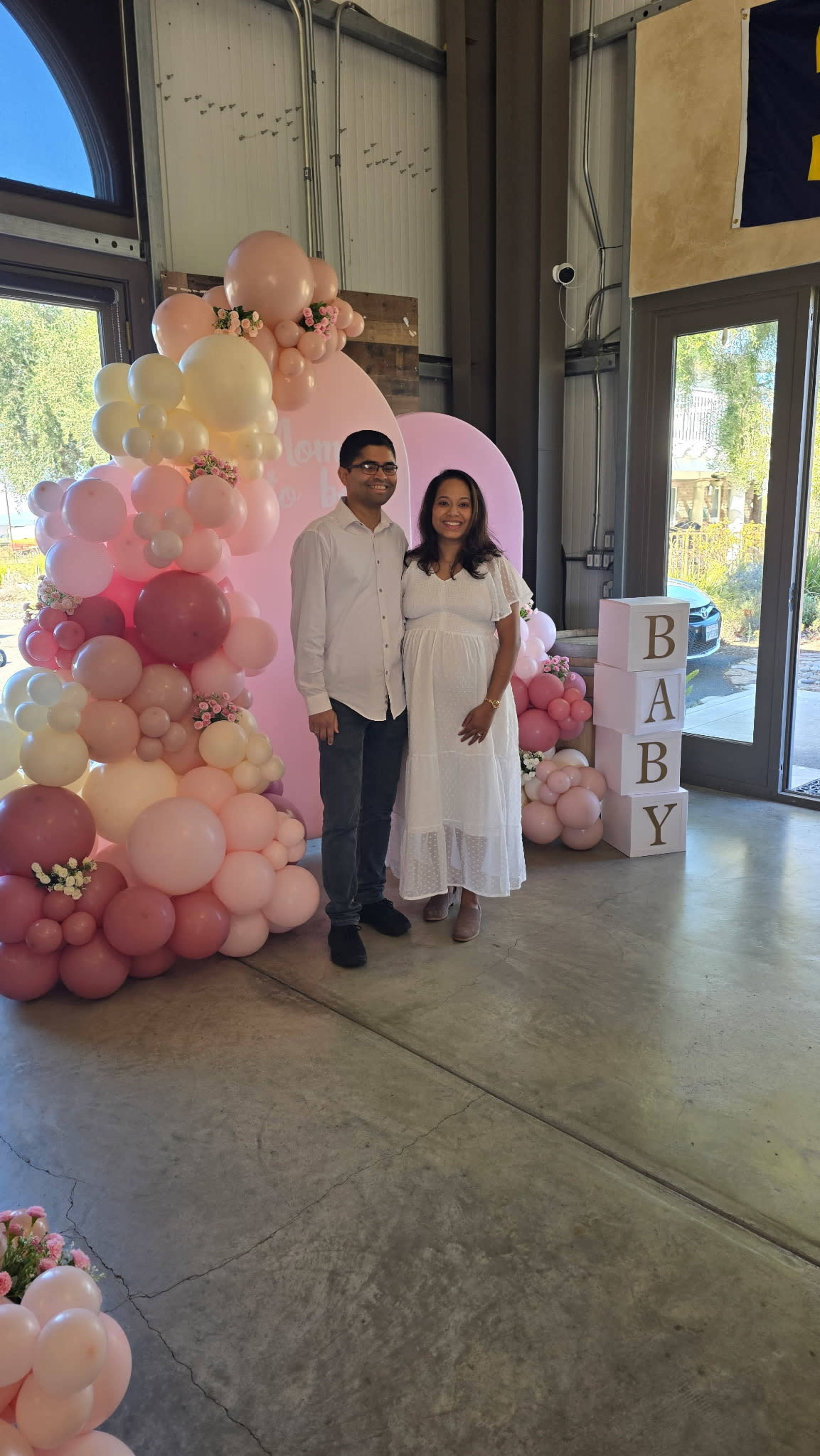 A couple stands in front of a balloon arch and decorative blocks spelling "BABY" at a celebration event.