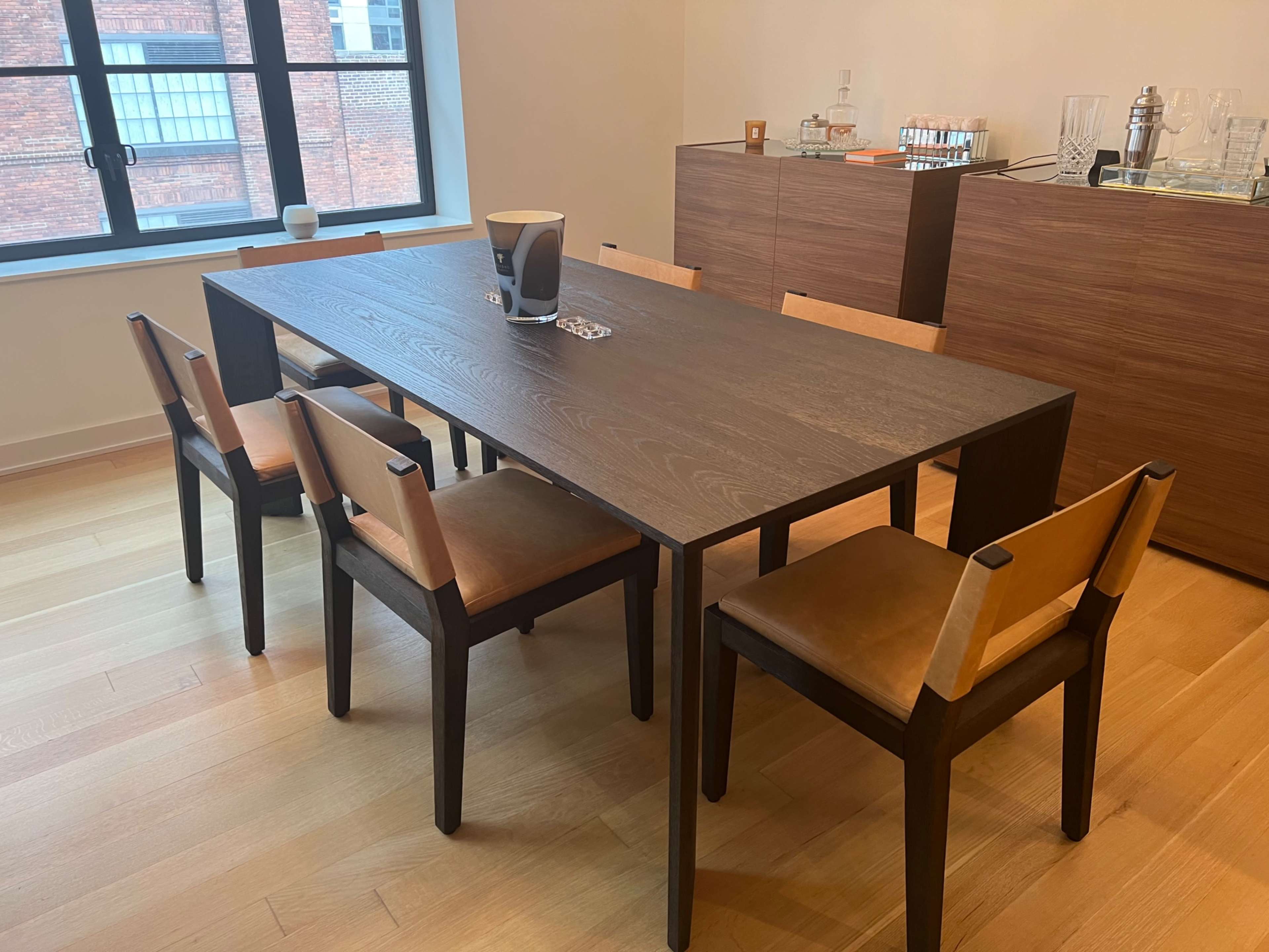 A wooden dining table with six chairs is set up in a well-lit room featuring a wooden cabinet and a large window.