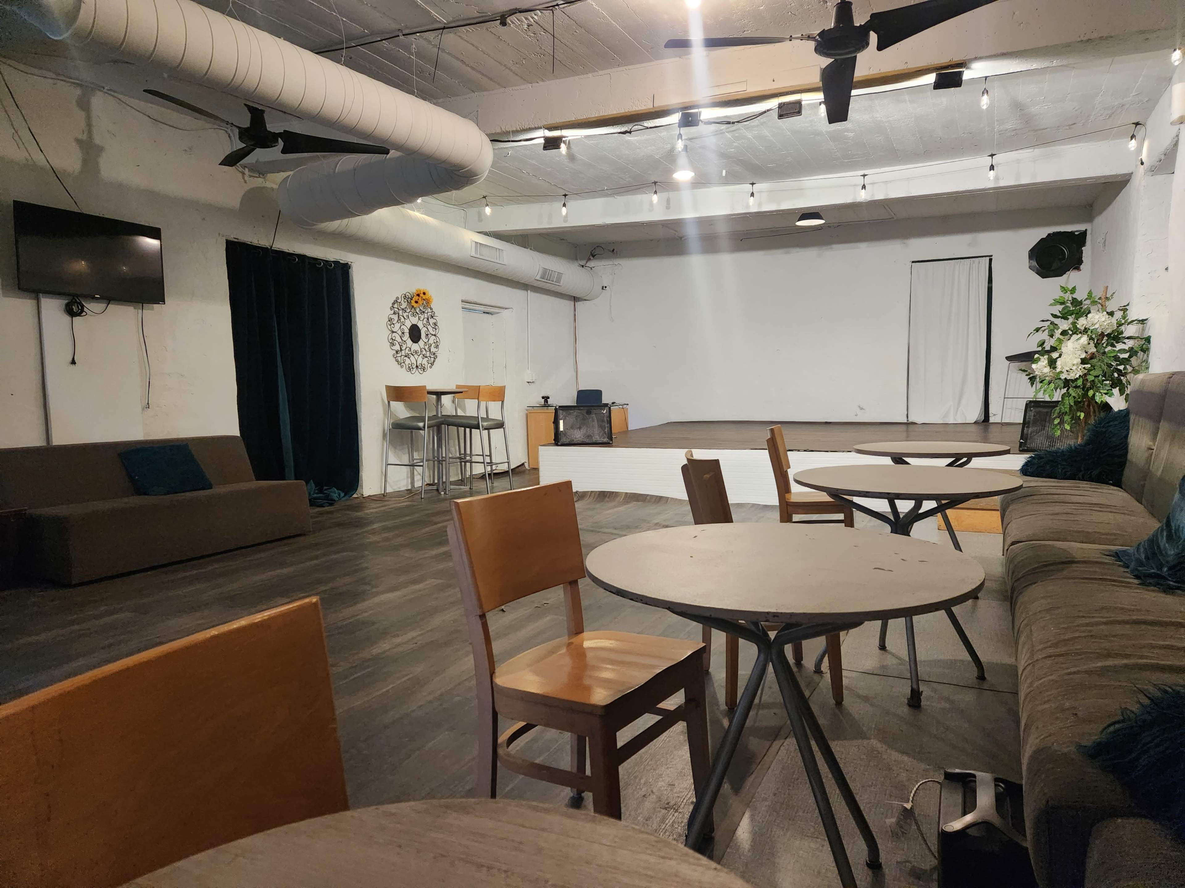 An empty room with a stage area, several tables and chairs, and a bar at the back.
