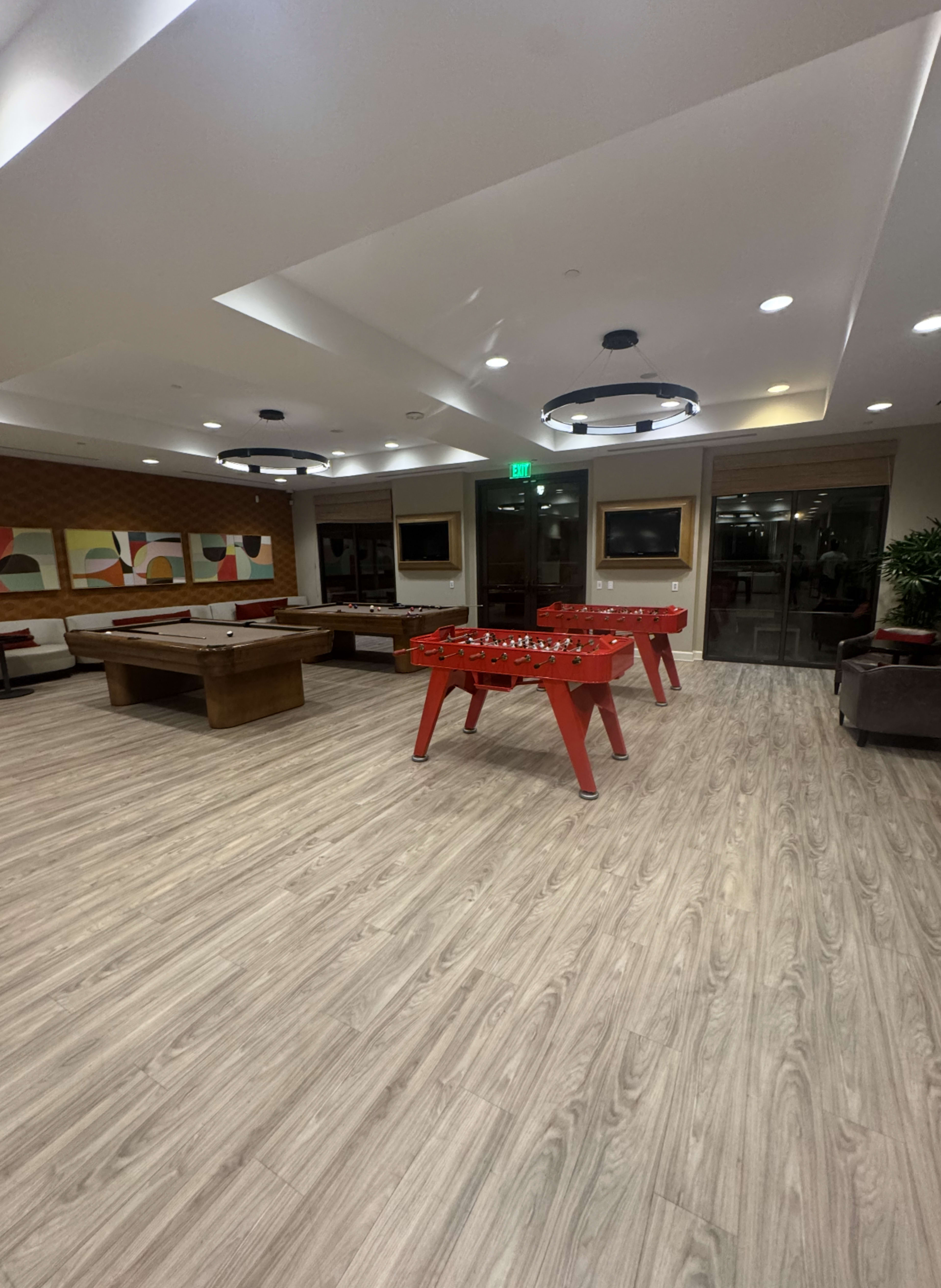 The image shows a recreational room with a pool table and foosball table on a wooden floor, surrounded by modern lighting and artwork on the walls.
