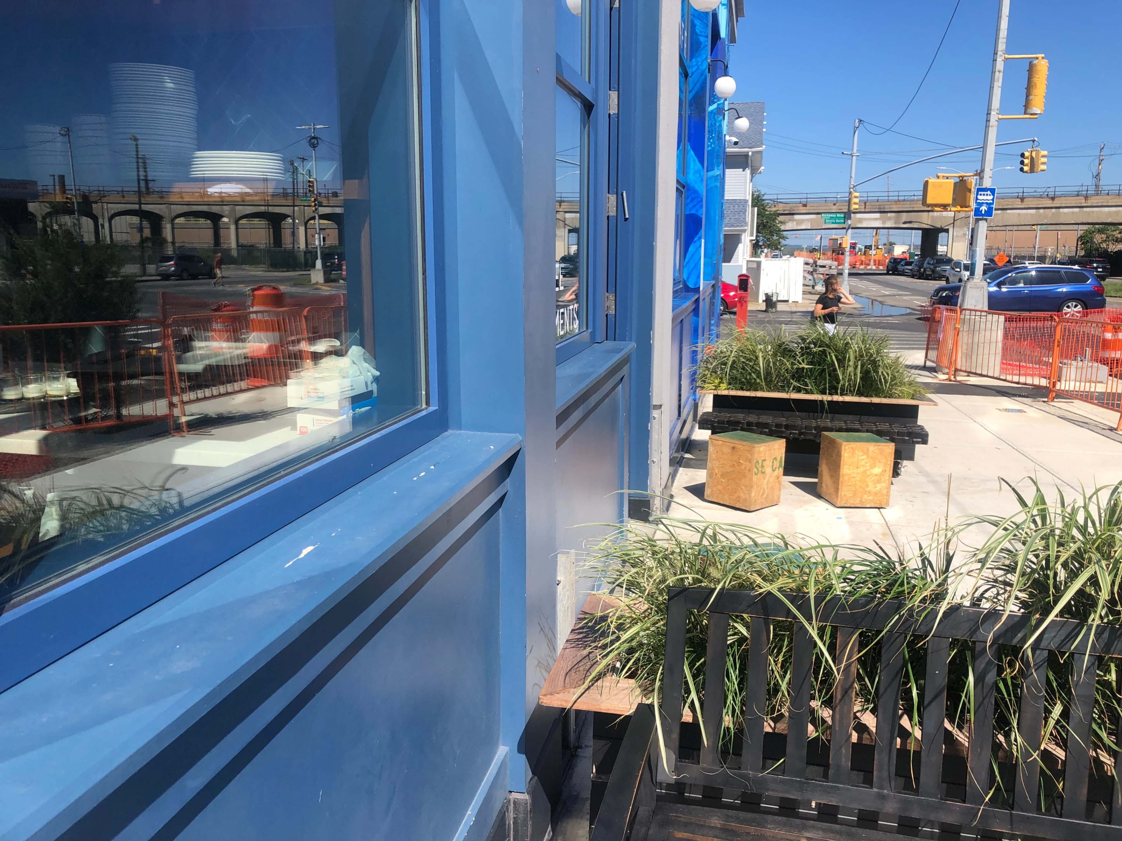 A blue building with large windows reflecting a city street and construction barriers, alongside planters containing grass.