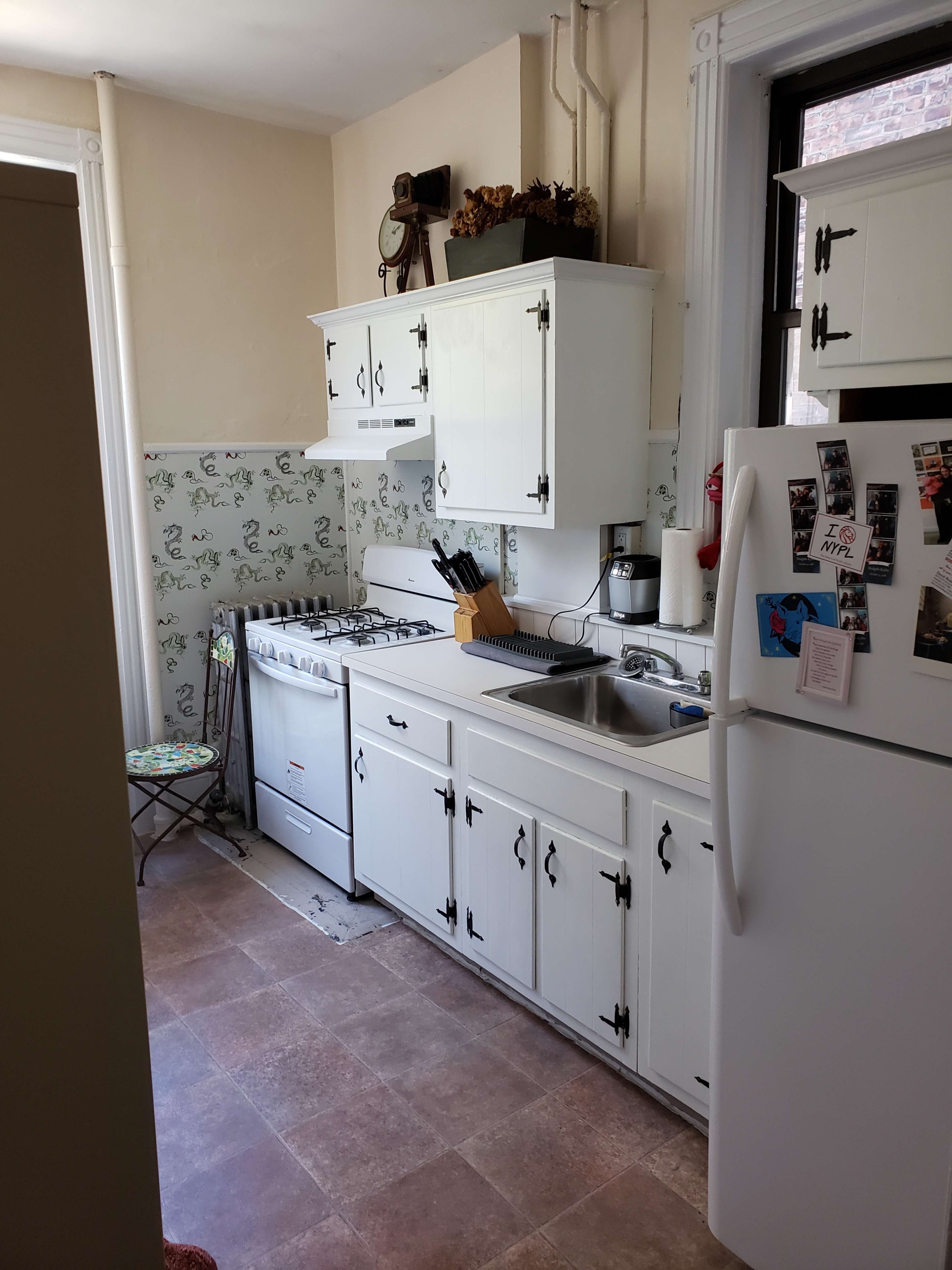 A small kitchen with white cabinets, a stove, sink, and refrigerator, along with patterned wallpaper and a tiled floor.