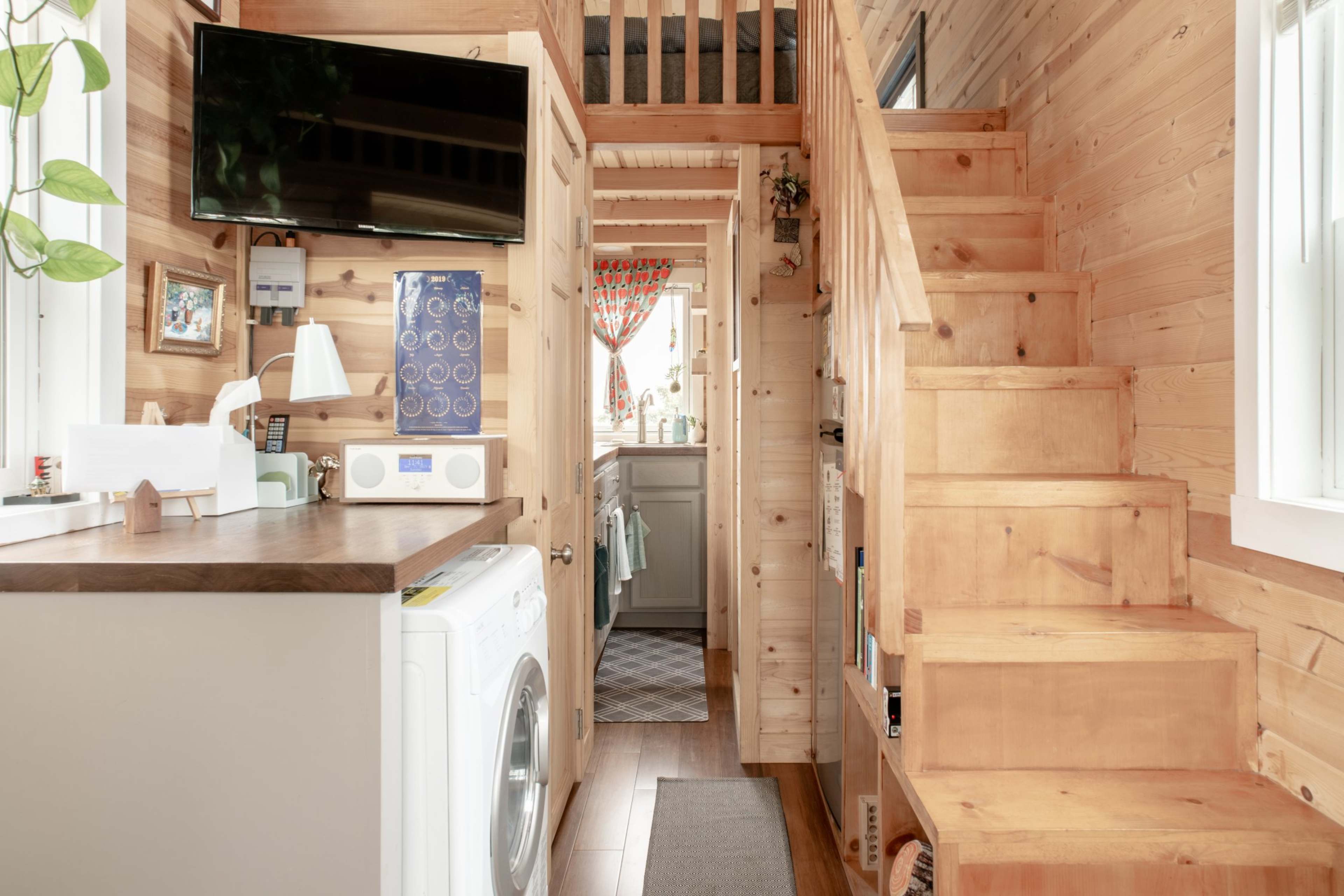 A wooden interior of a small home, featuring a staircase, a laundry area near a microwave, and a view of a kitchen in the background.