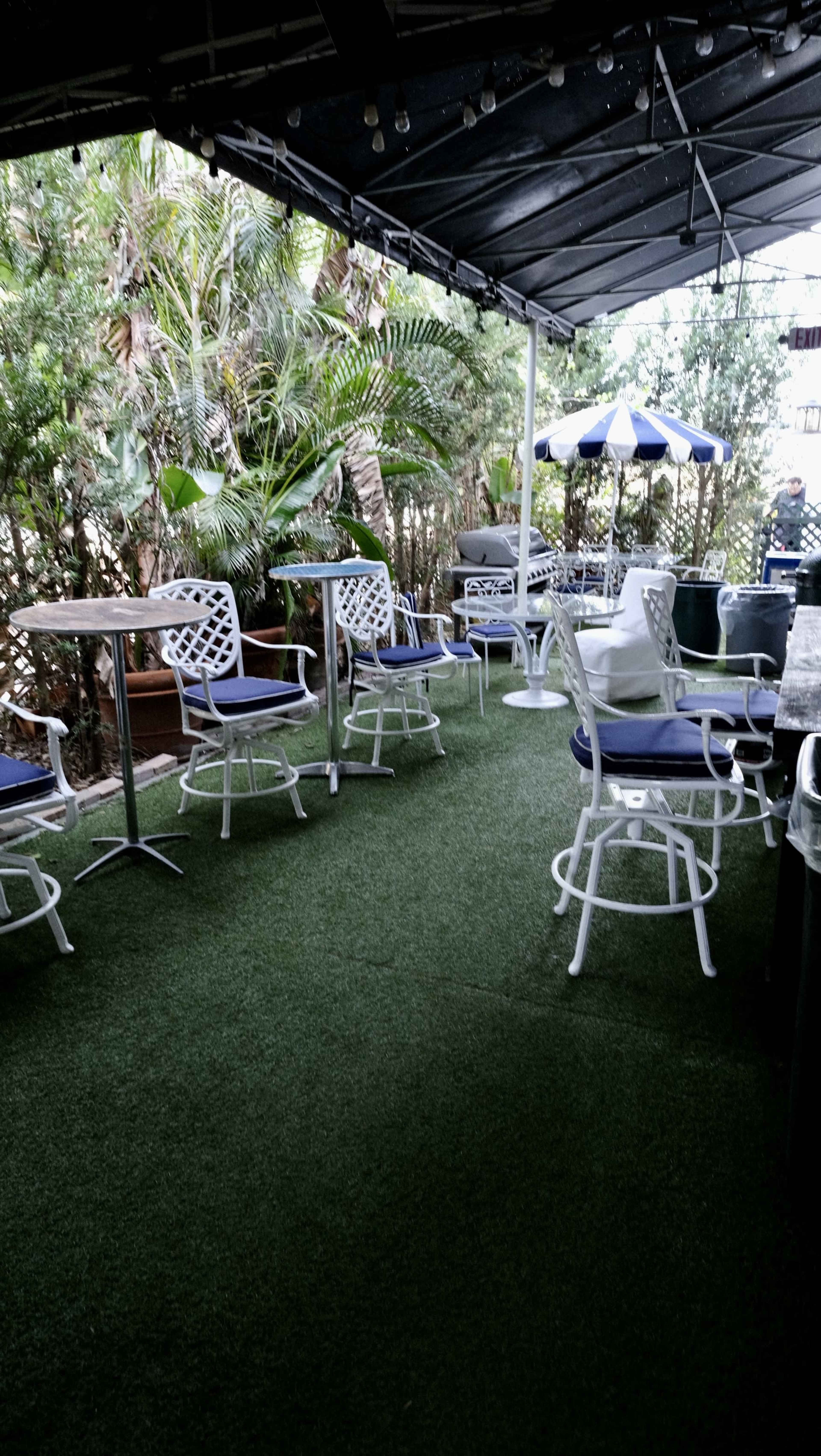 The image shows an outdoor patio with white metal furniture, including tables and chairs, set on artificial grass surrounded by greenery.