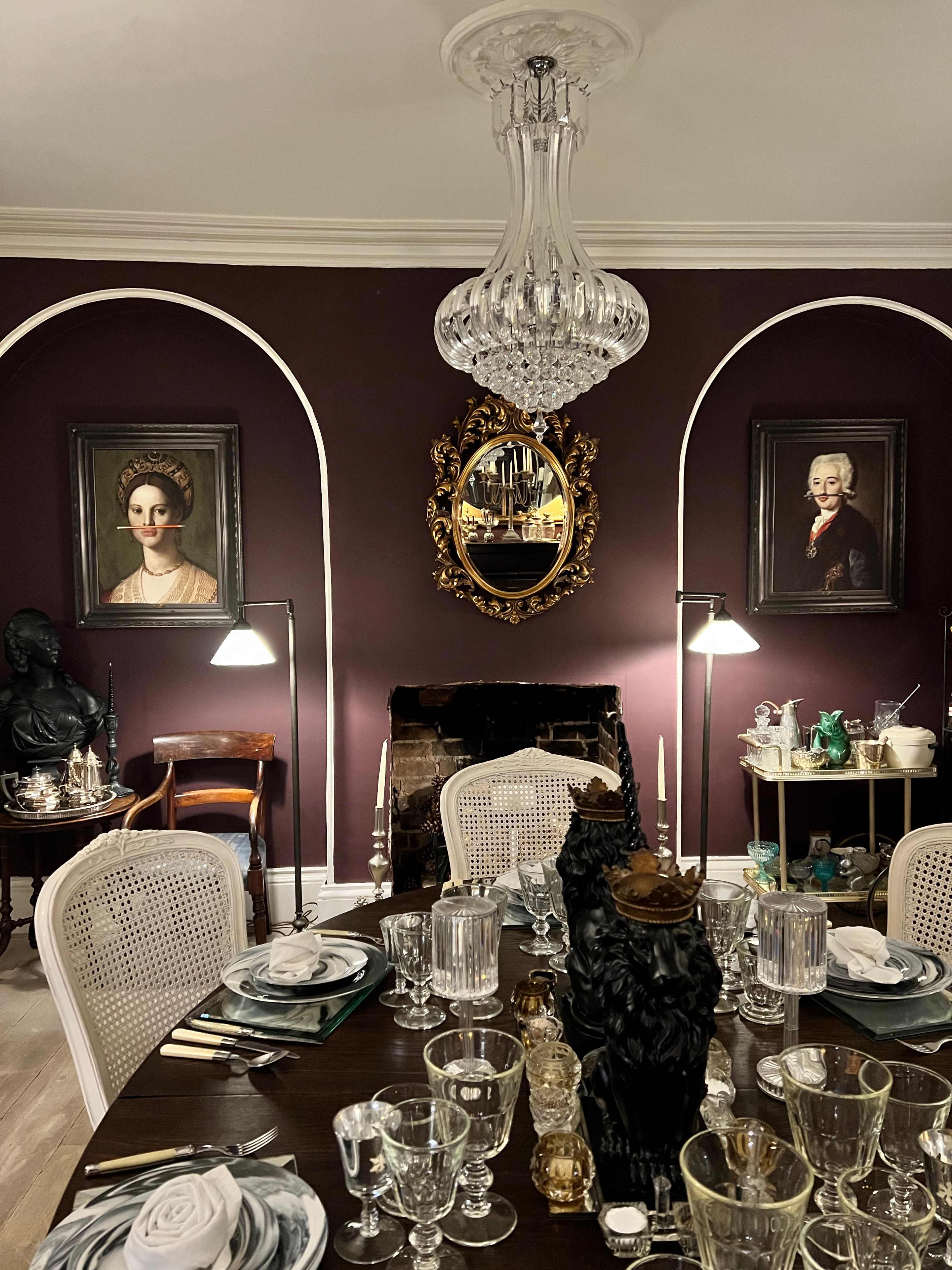 The elegantly set dining room features a dark purple wall adorned with two framed portraits, a large chandelier, and a table arranged with various glassware and decorative items.