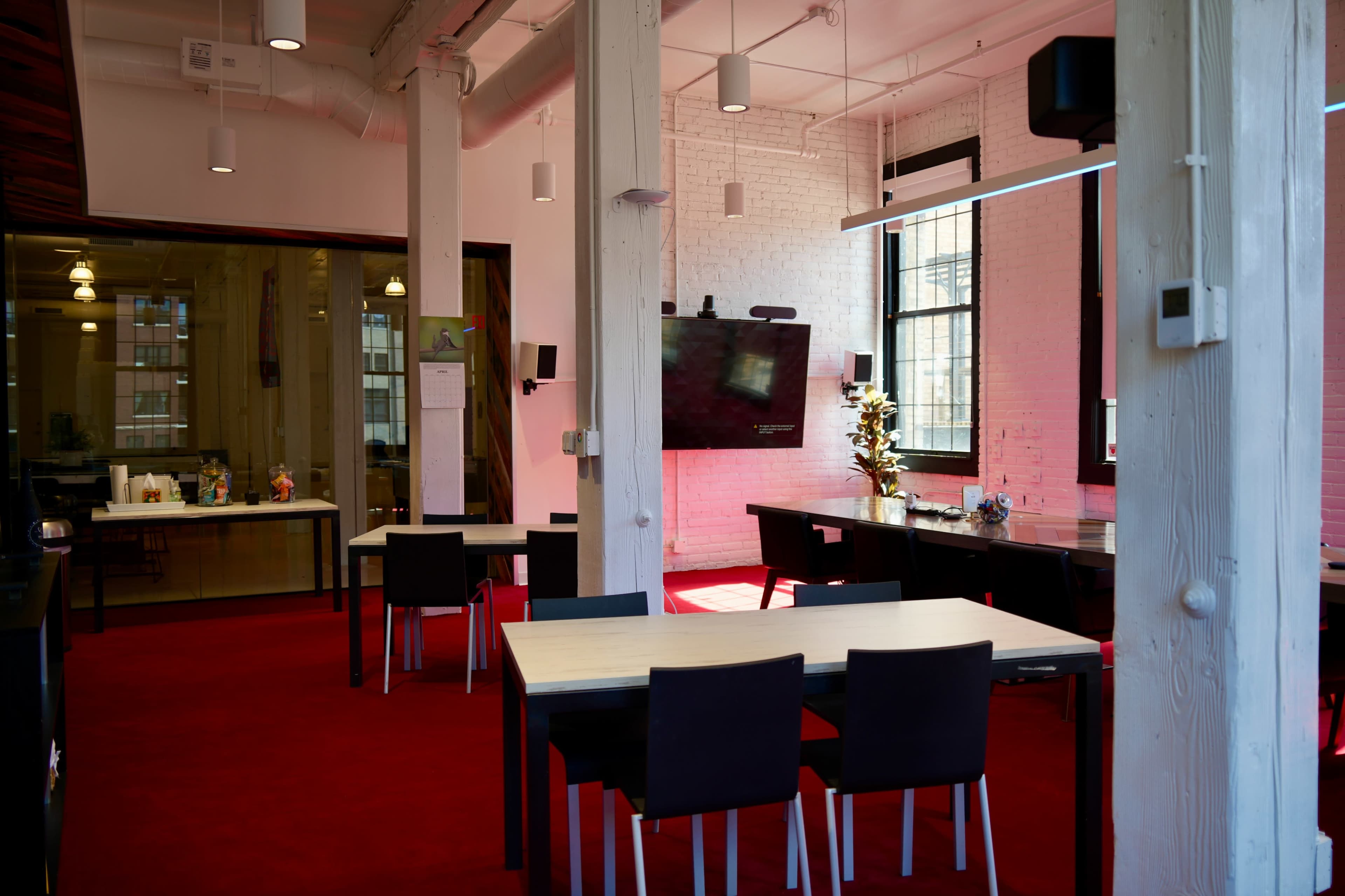 The image shows a modern workspace with tables and chairs arranged on a red carpet, featuring large windows and a video screen mounted on the wall.